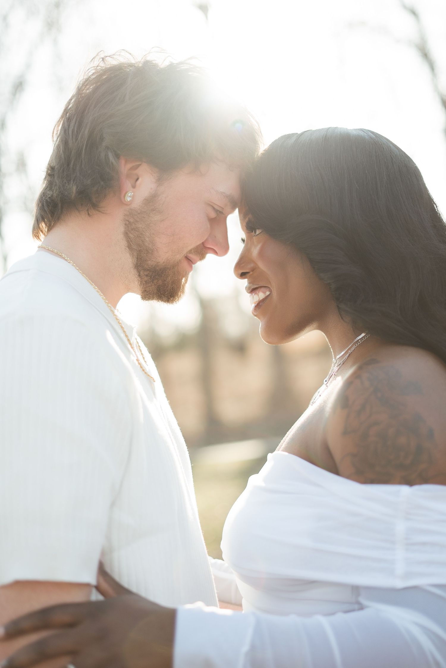 Interracial couple in white outfits sharing a romantic moment outdoors with warm backlit sunlight.