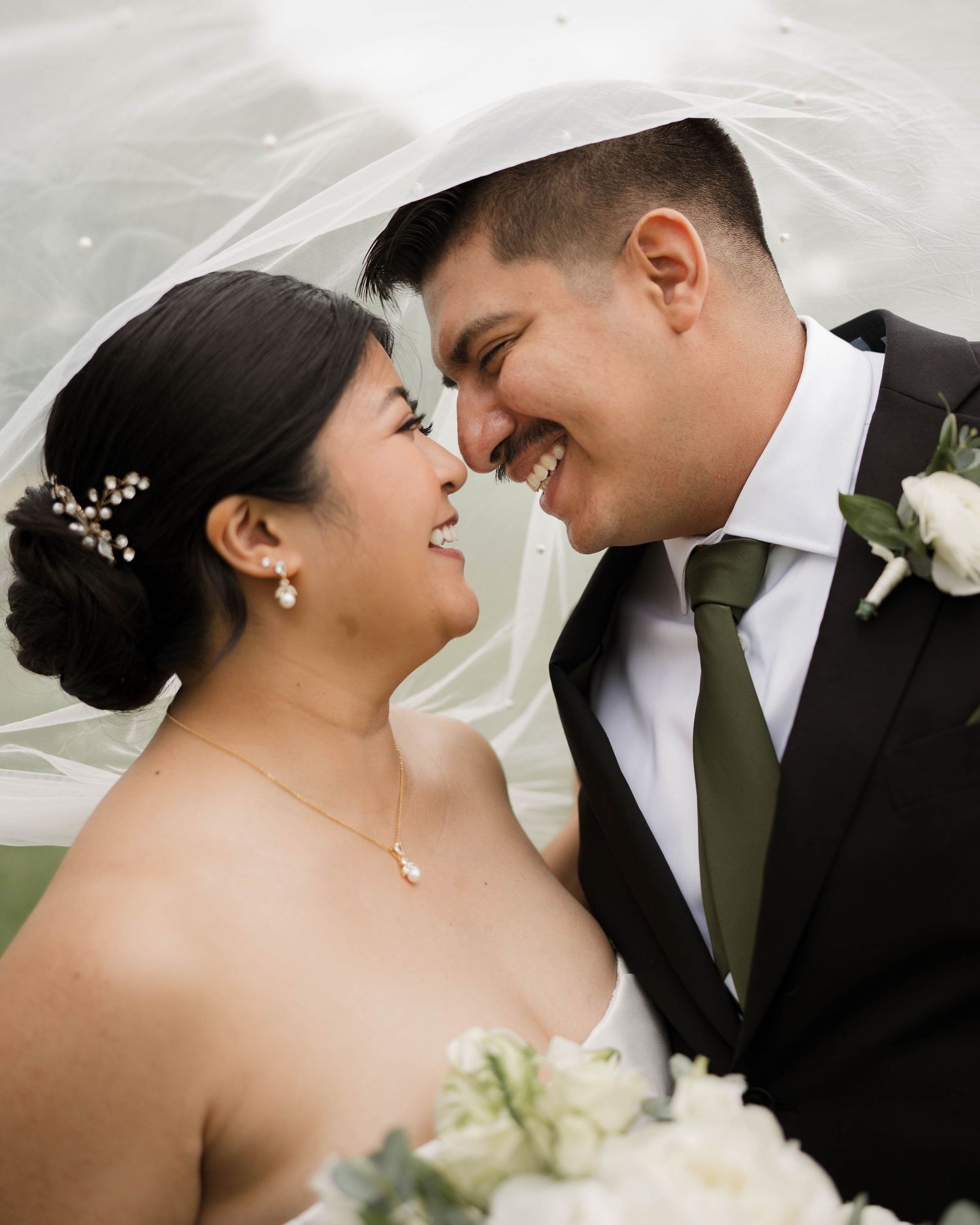 Bride and groom smiling at each other under a veil, bride holding white bouquet, groom in black suit with green tie.