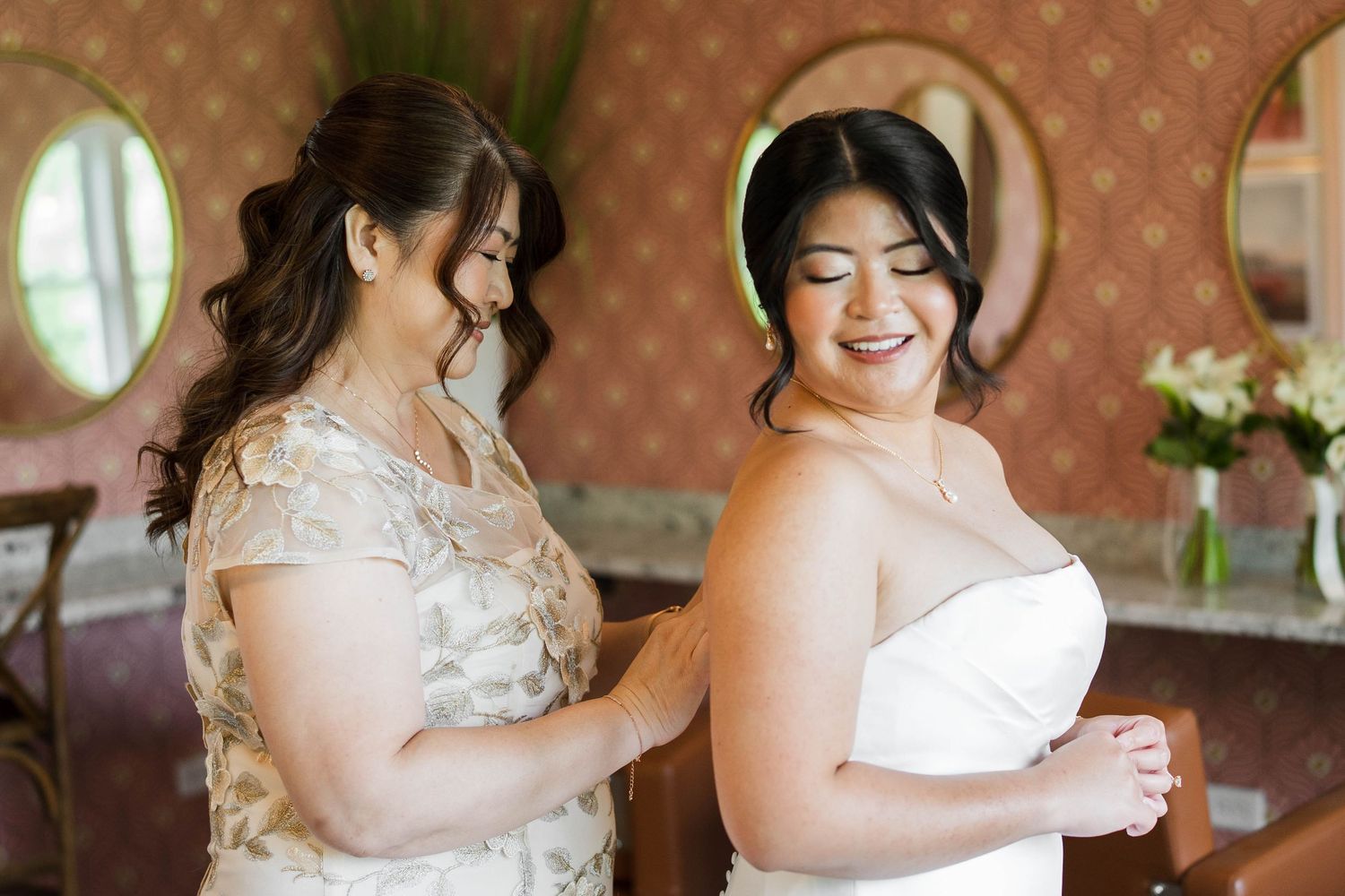 A bride in a white gown smiles as her mother helps fasten her dress in an elegant rose-wallpapered bridal suite.