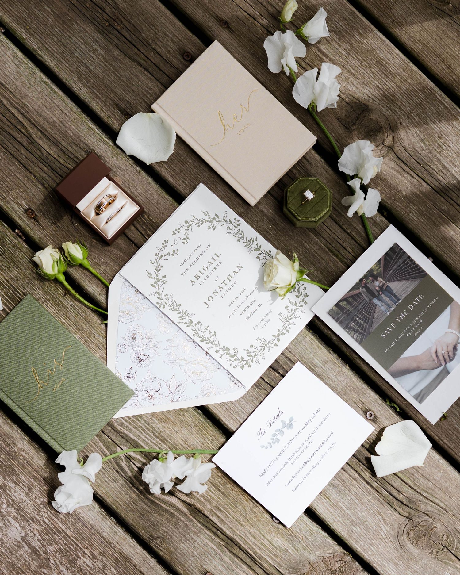 Flat lay of sage green and gold wedding invitation suite with rings and white florals on wood.