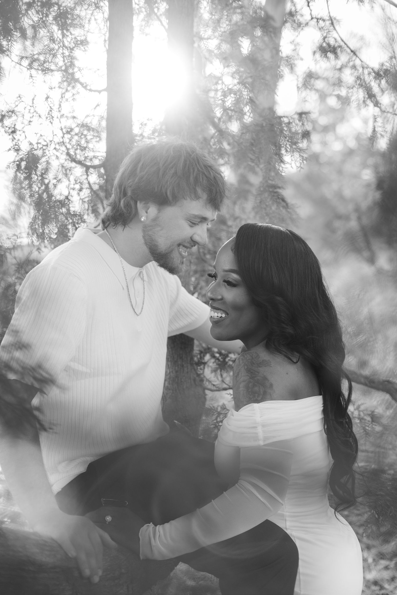 Black and white photo of a smiling interracial couple sharing a joyful moment outdoors among trees in soft sunlight.