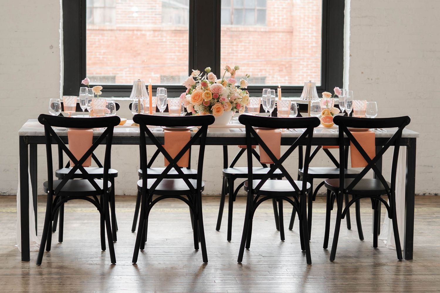 Elegant wedding reception table with black cross-back chairs, peach napkins, floral centerpiece, and large windows in background.