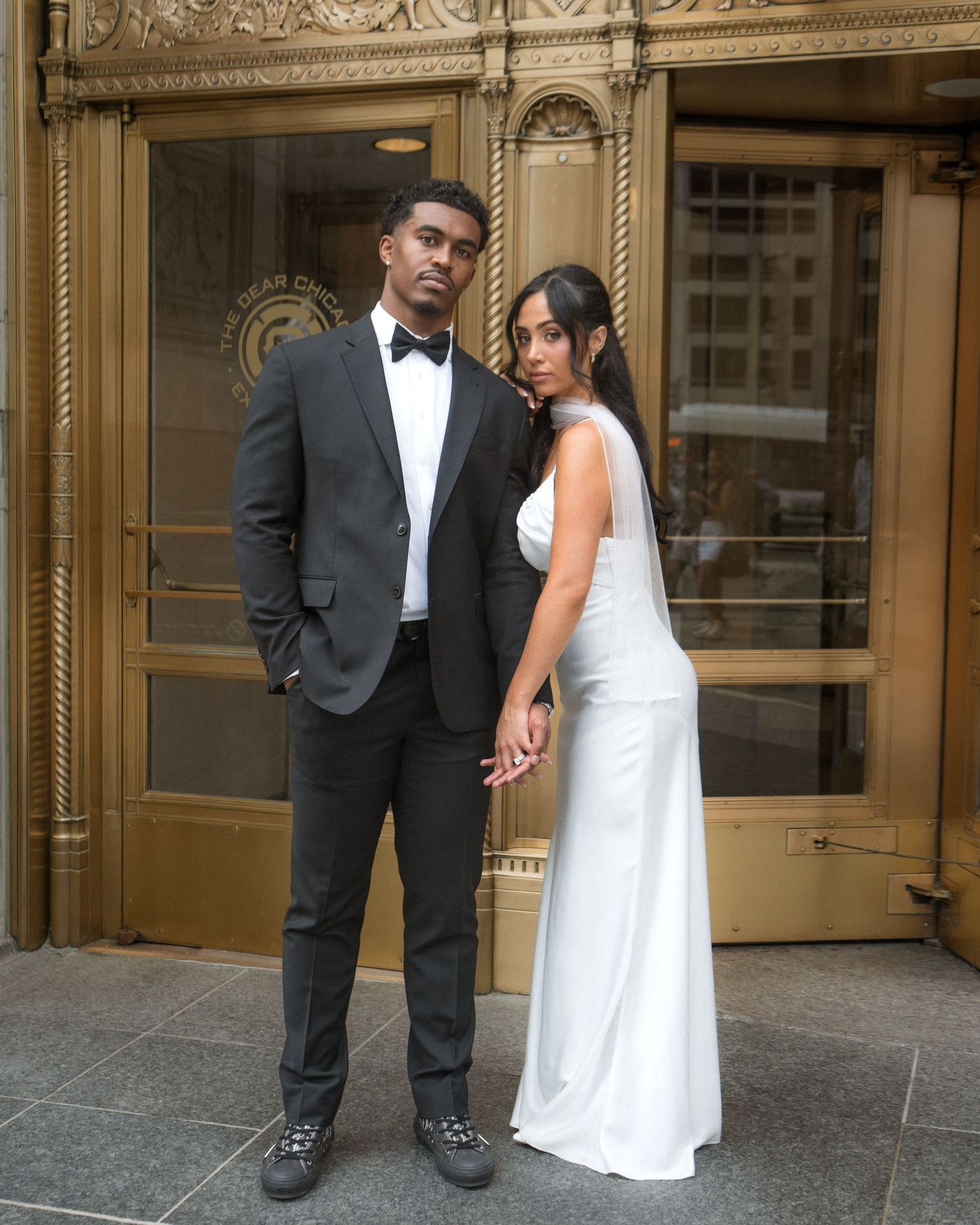 Elegant couple in wedding attire pose outside a grand building, groom in black tuxedo and bride in white gown.