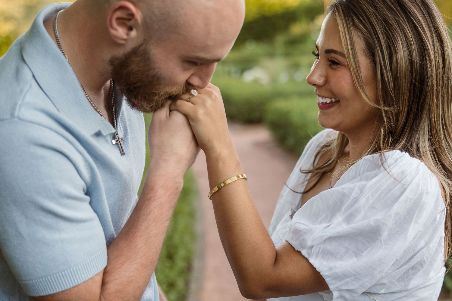 A man gently kisses a woman's hand while she smiles warmly during a romantic outdoor engagement photo session.