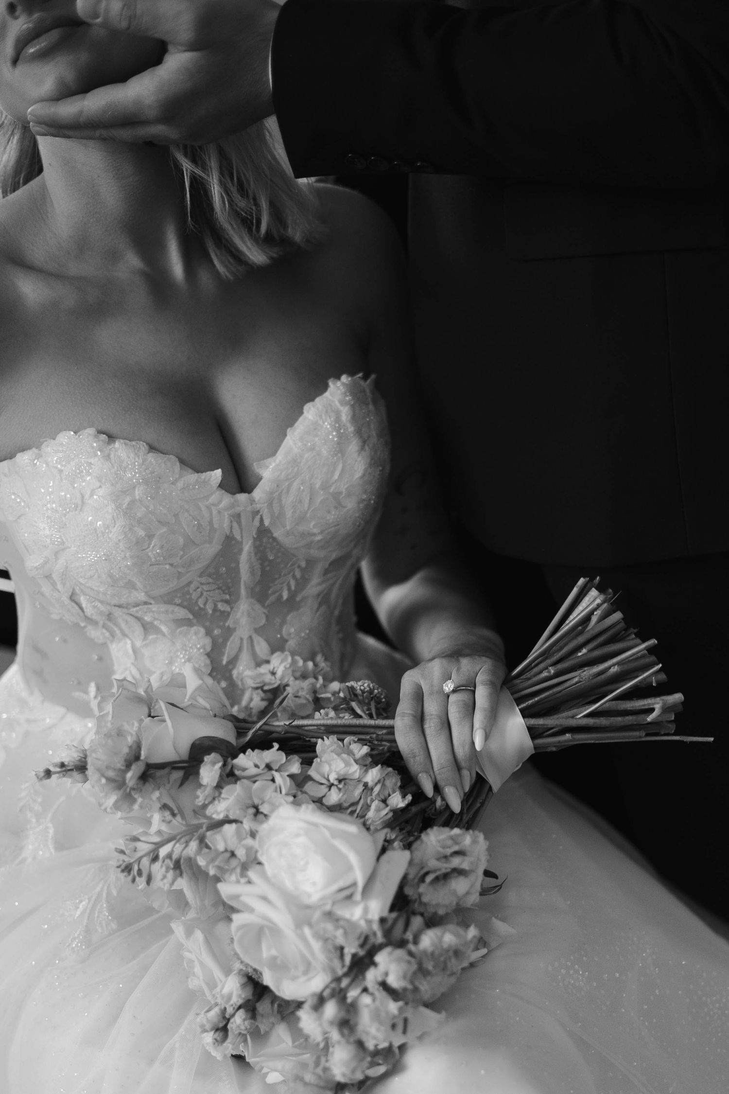 Black and white photo of a bride in a strapless lace gown holding a floral bouquet while embraced by her groom.