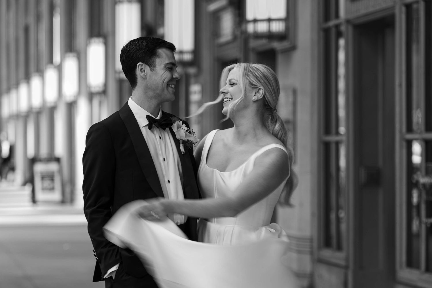 Black and white photo of a smiling bride and groom in formal attire sharing a joyful moment outside a building.