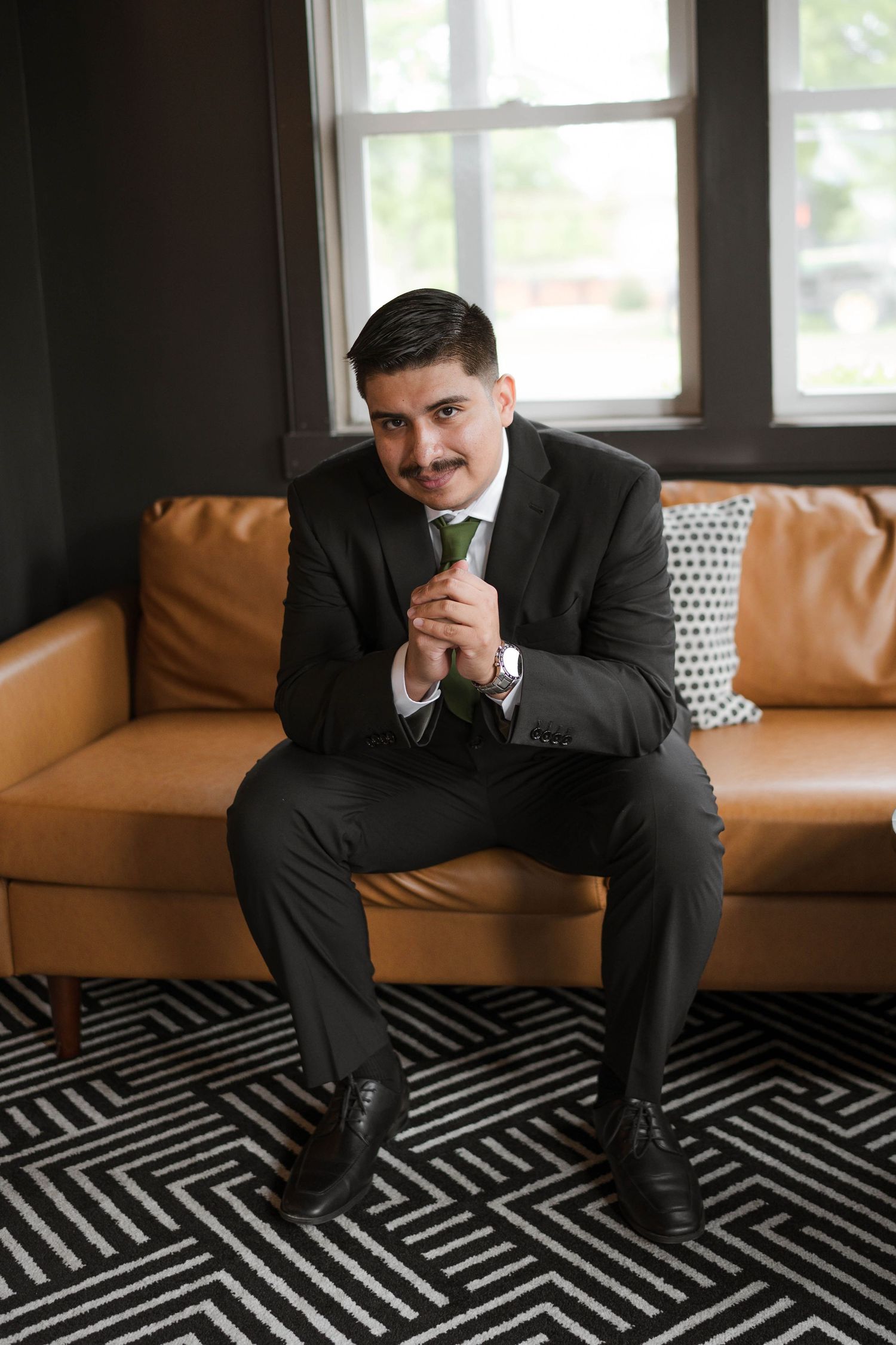 Groom in dark suit with green tie sitting on a leather couch before the wedding ceremony.