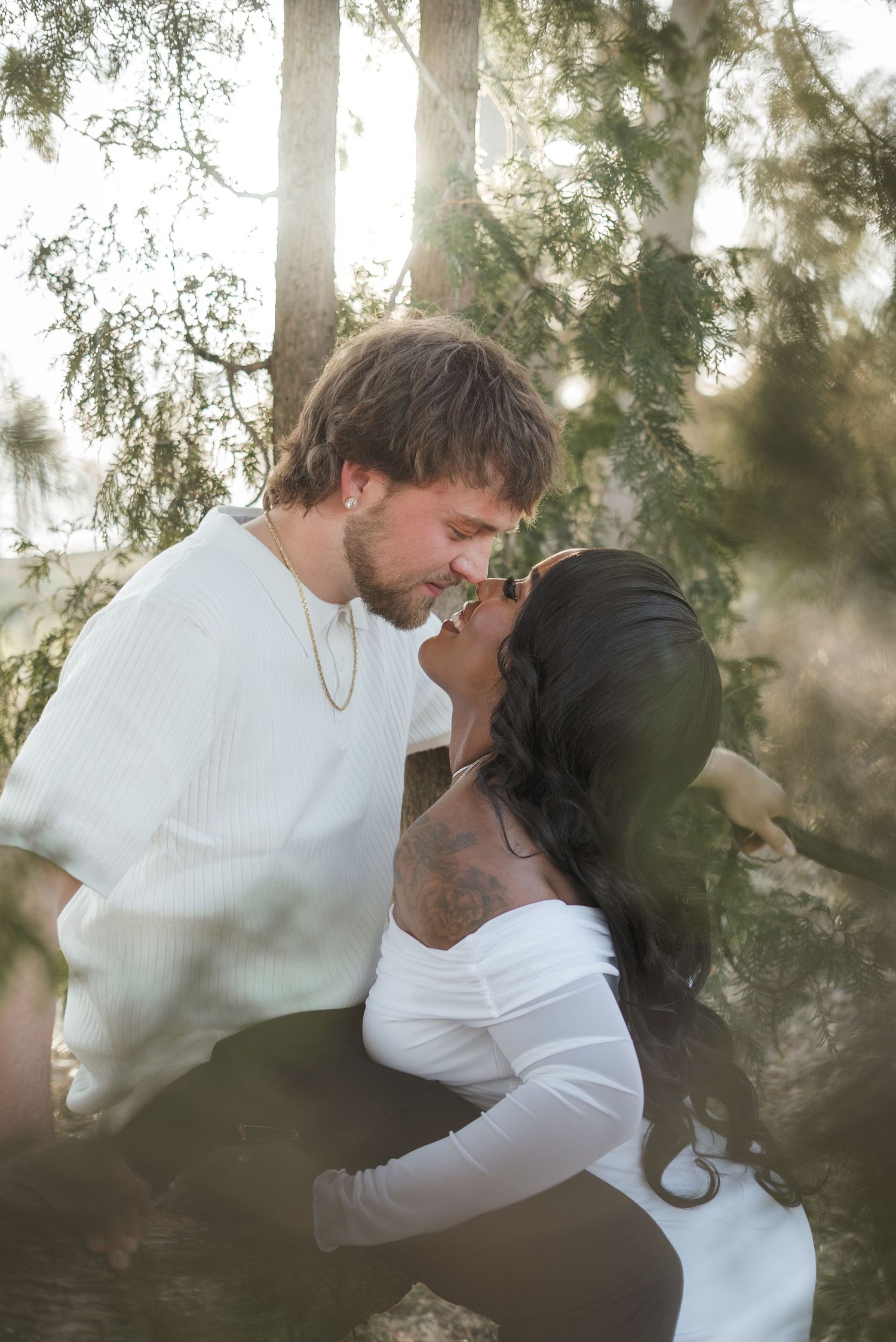 A couple shares a romantic almost-kiss moment outdoors, surrounded by sunlit trees, both dressed in white outfits.