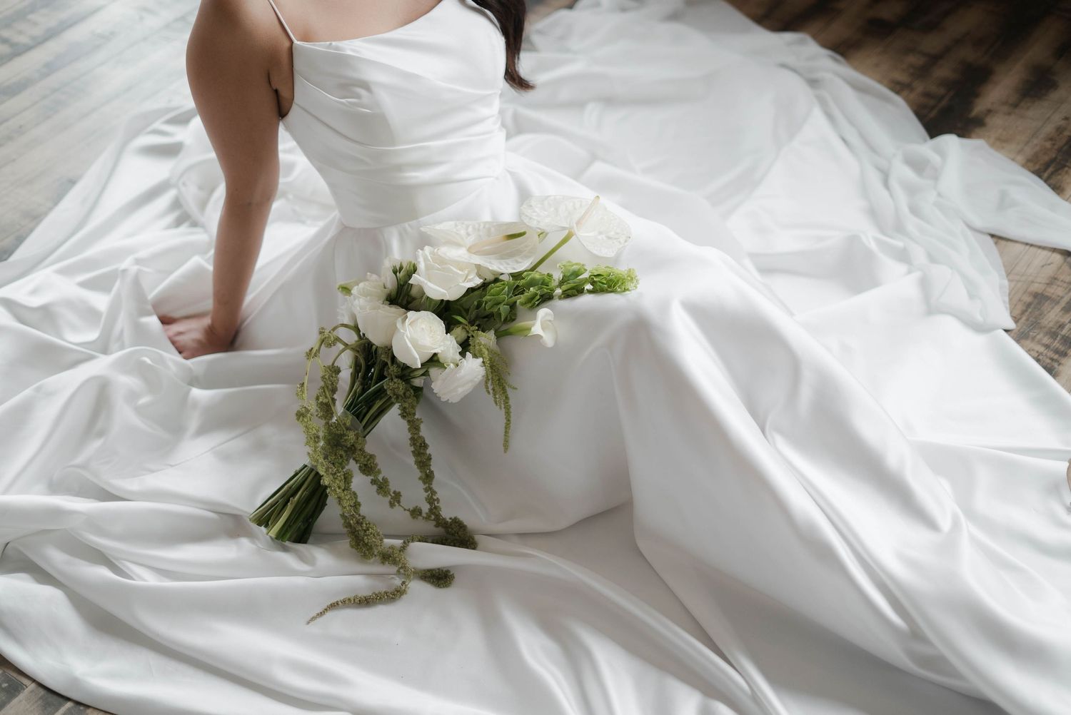Bride in white satin wedding gown sitting on floor with elegant white floral bouquet draped across the dress train.
