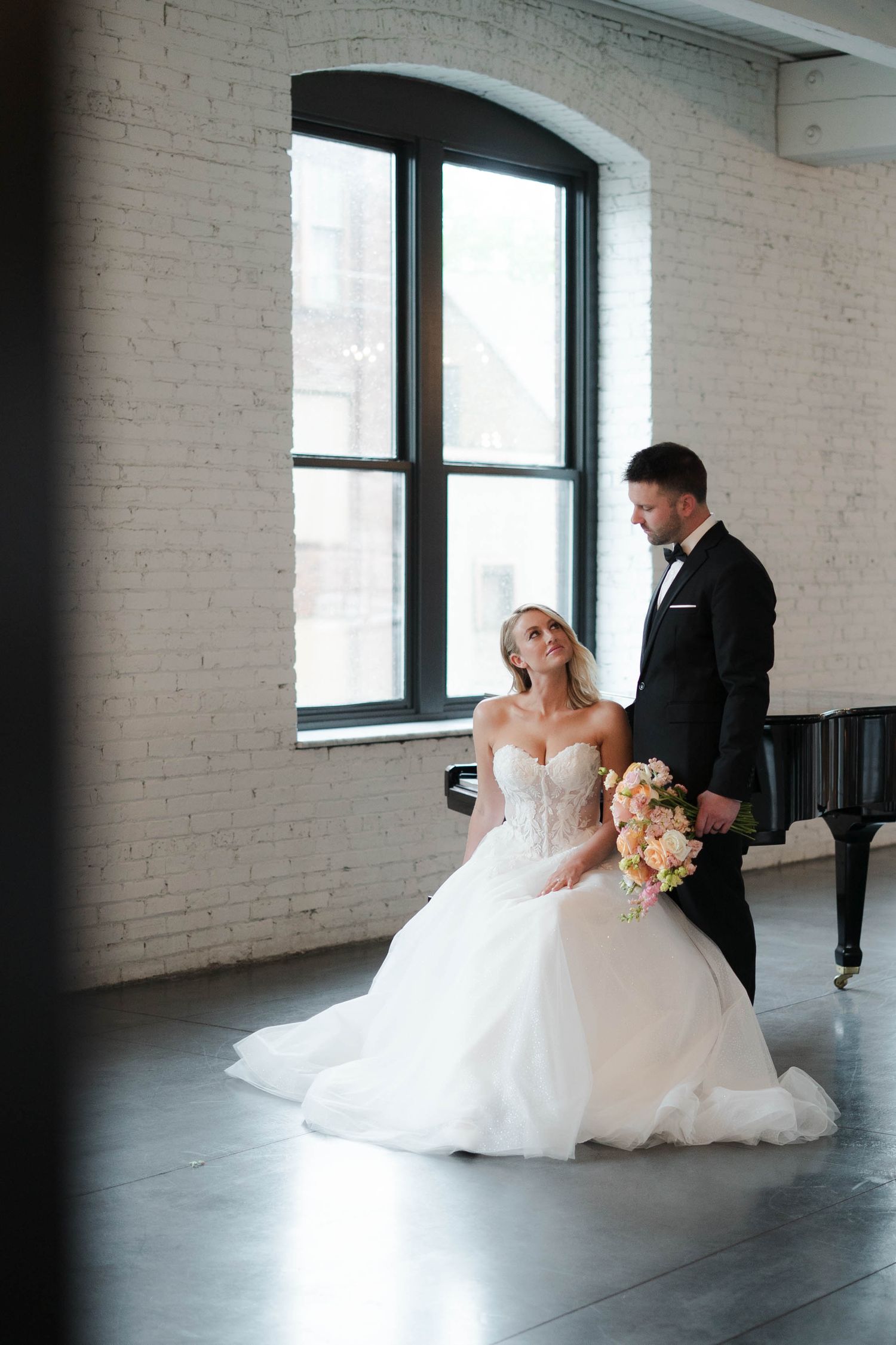 Bride in white gown sits holding bouquet while groom in tuxedo stands beside her near arched window.
