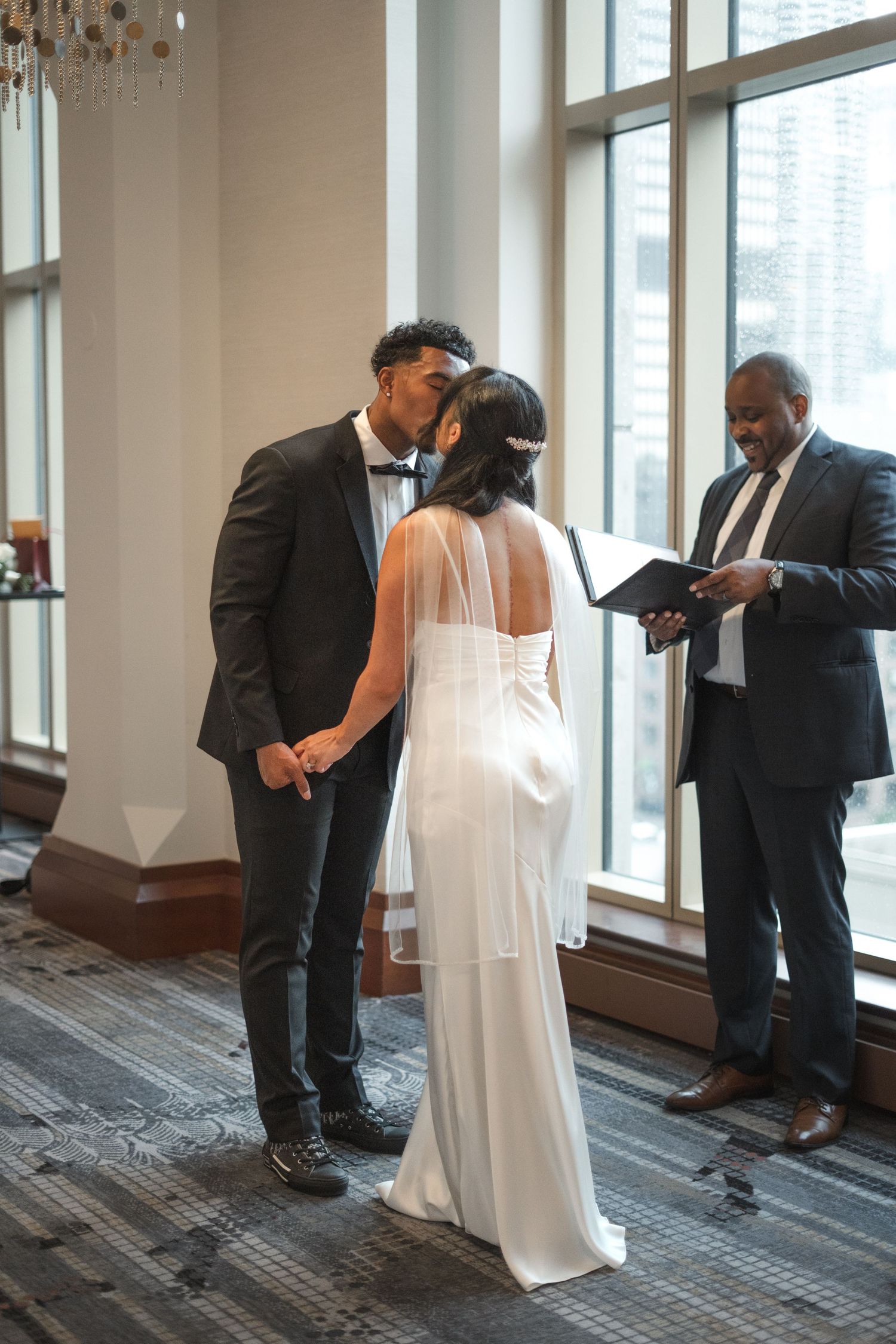 Bride and groom share first kiss during indoor wedding ceremony while officiant reads from book near large windows.