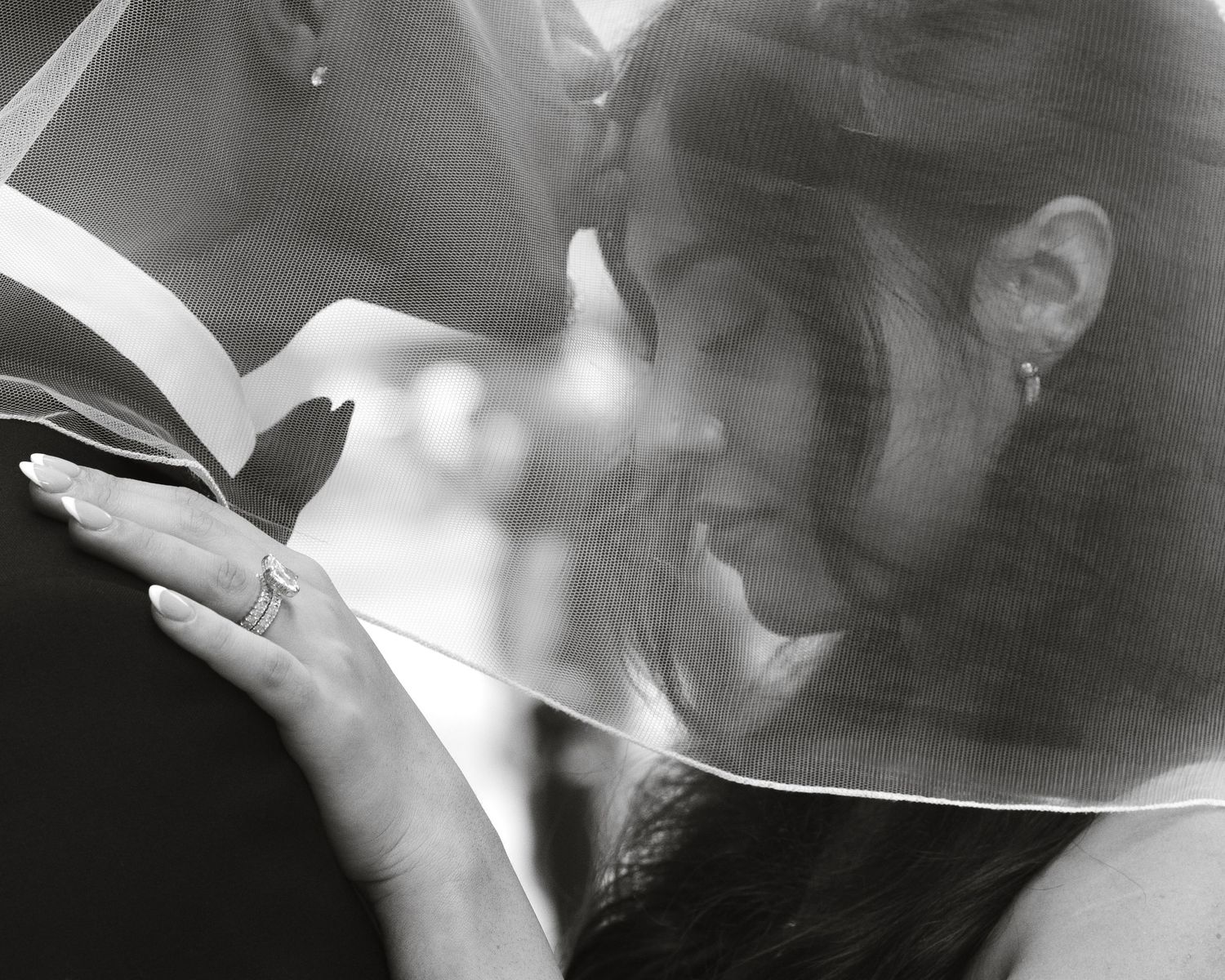 Black and white wedding photo of bride and groom sharing a kiss under a veil, showcasing a diamond ring.