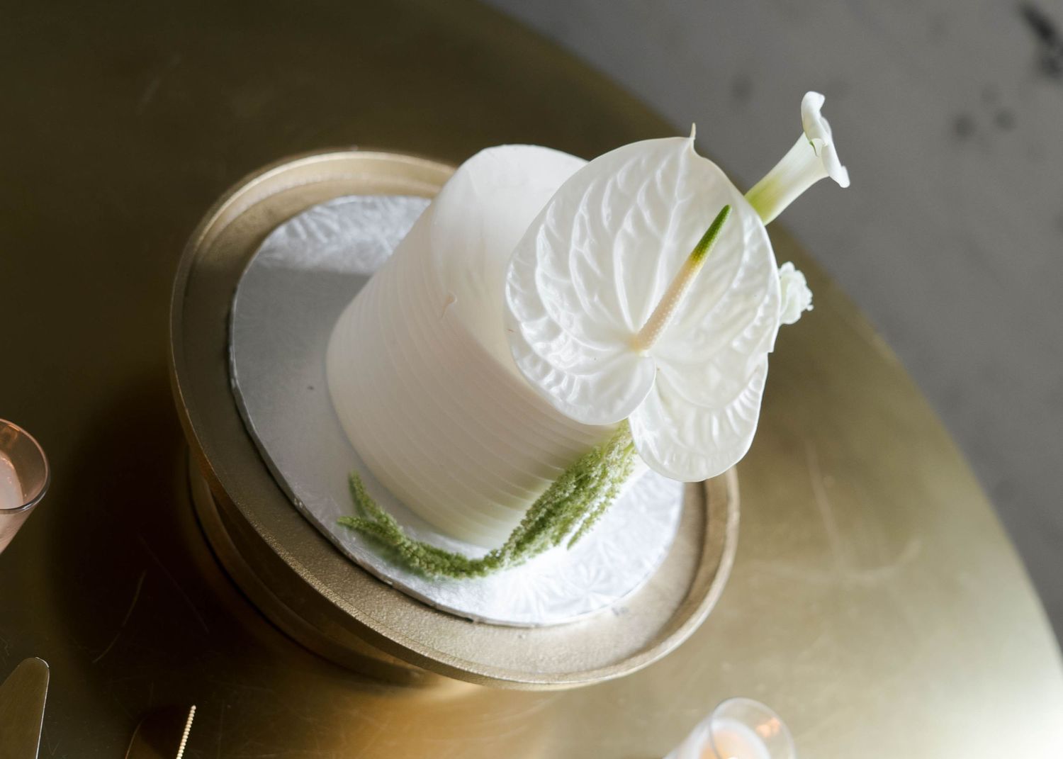 Elegant two-tier white wedding cake topped with white anthurium and calla lily on a gold stand.