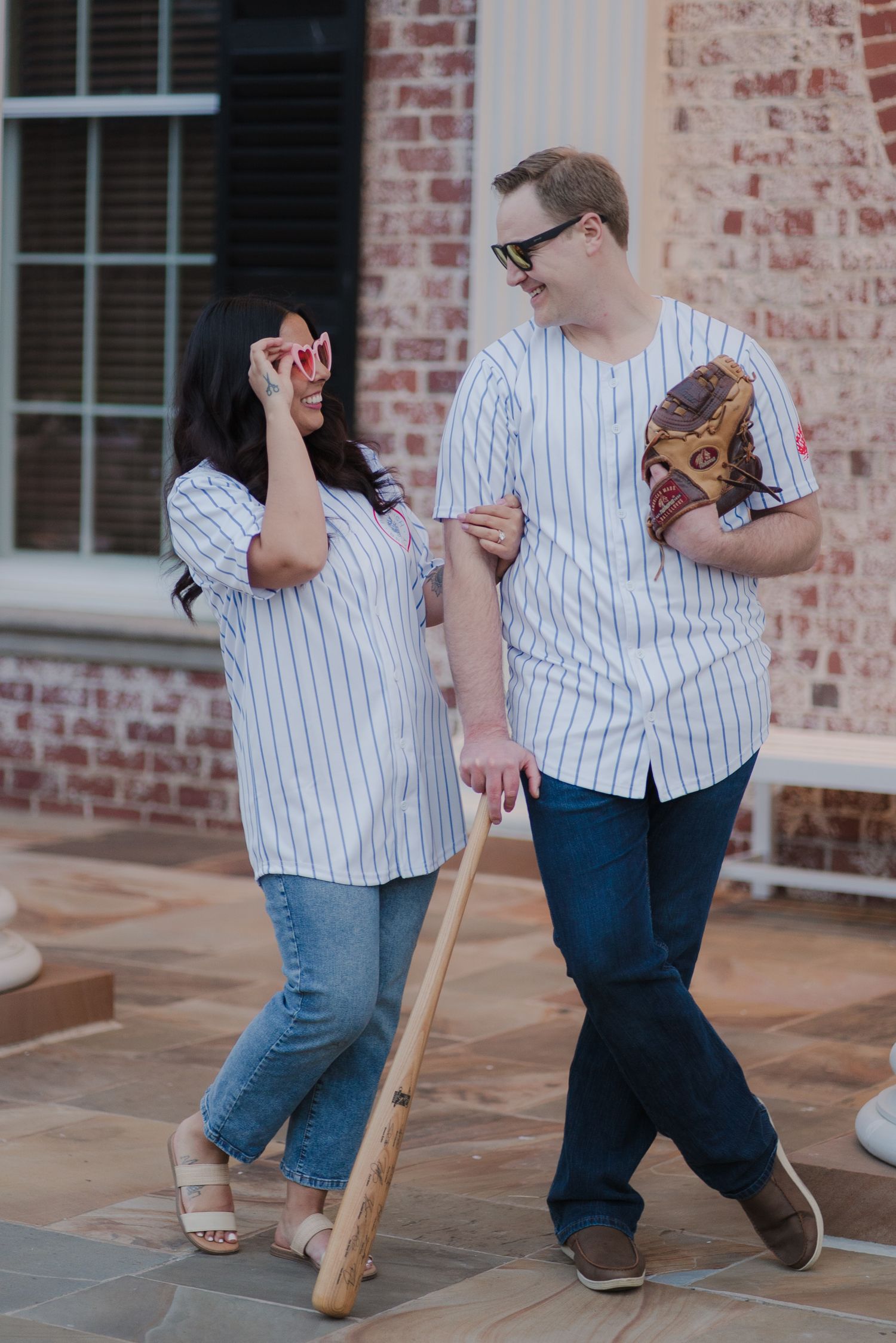 Couple in matching pinstripe baseball jerseys walking together with bat and glove, laughing outdoors.