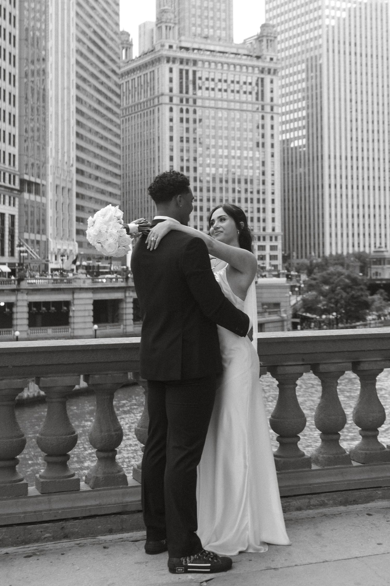 Black and white photo of a bride and groom embracing on a bridge with Chicago city skyscrapers in the background.