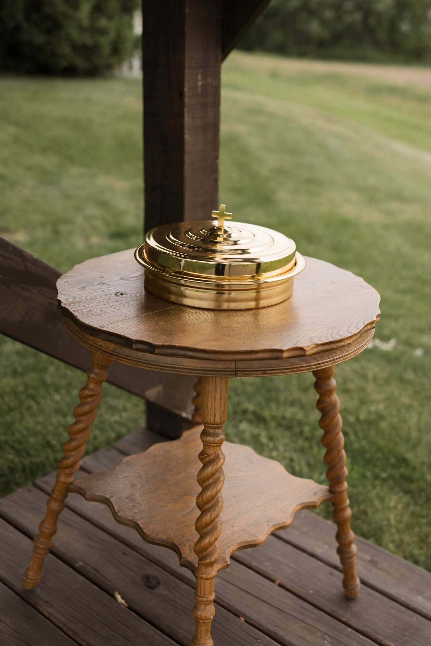 Antique wooden three-legged side table with ornate twisted legs holding a gold communion plate on a rustic wooden deck.