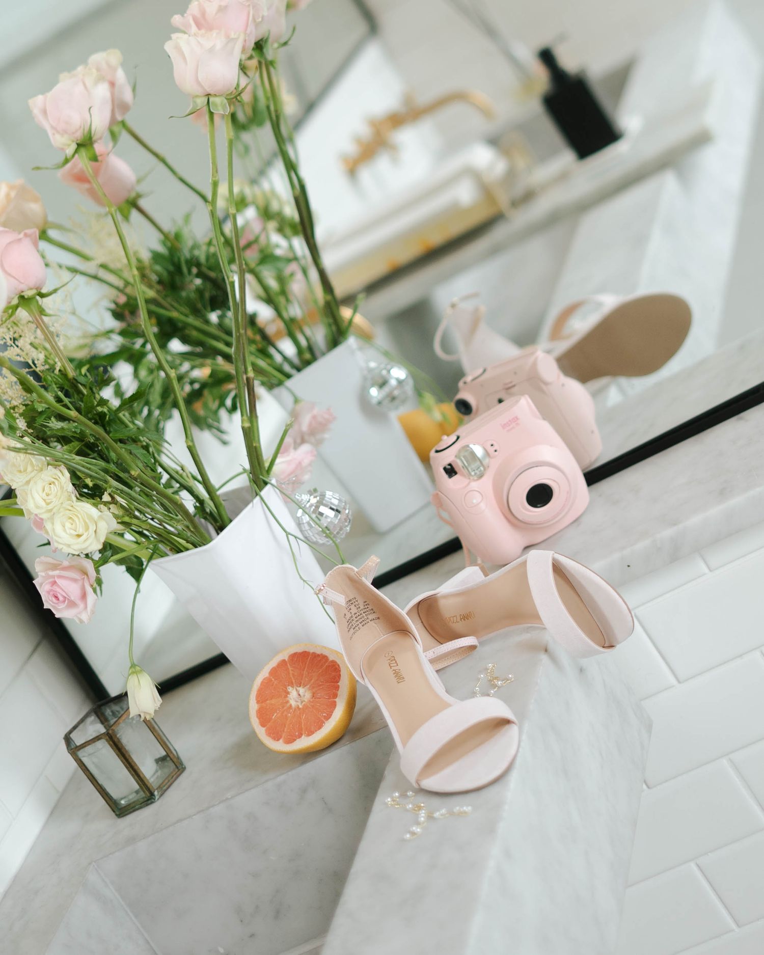 Pink Instax camera, white heeled sandals, grapefruit, and pink roses in a vase on a marble bathroom counter.