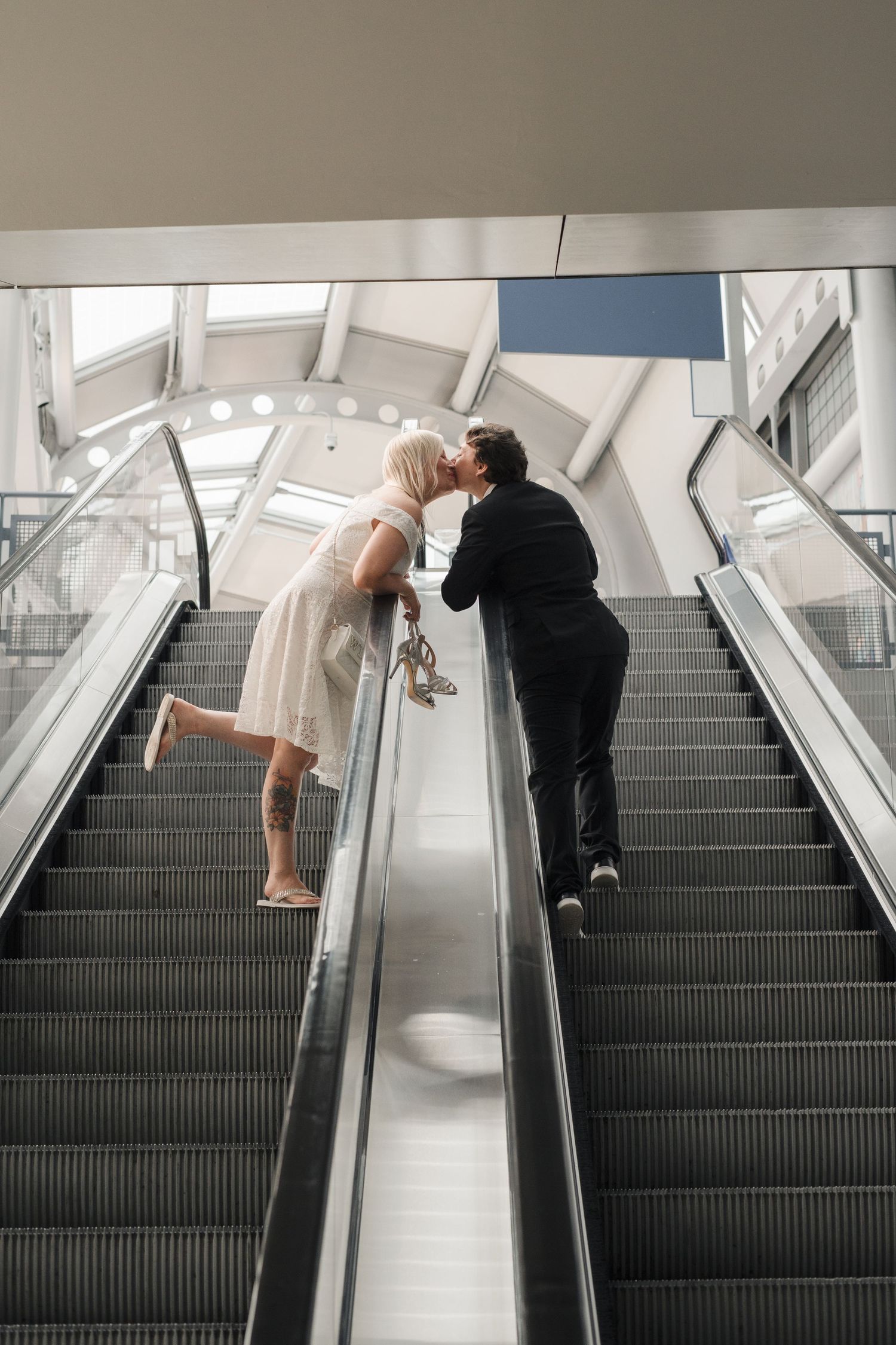 Couple sharing a romantic kiss on an escalator, woman in white dress holding heels, man in black suit.