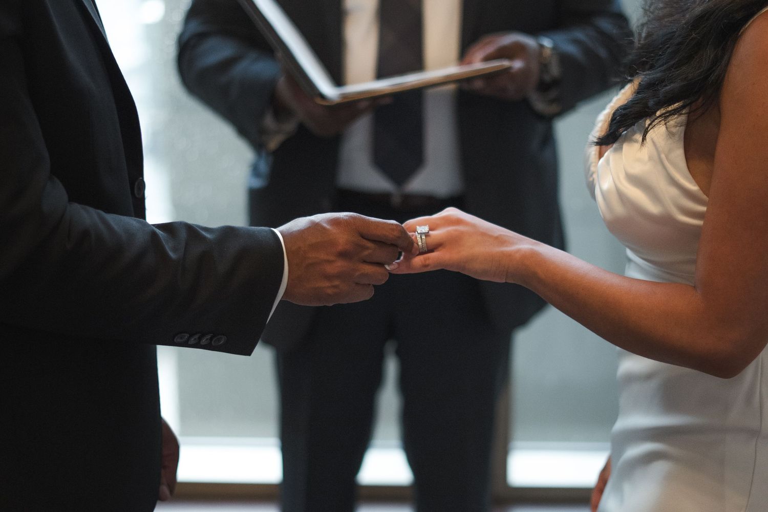 Groom placing a wedding ring on bride's finger during ceremony, with officiant holding book in background.