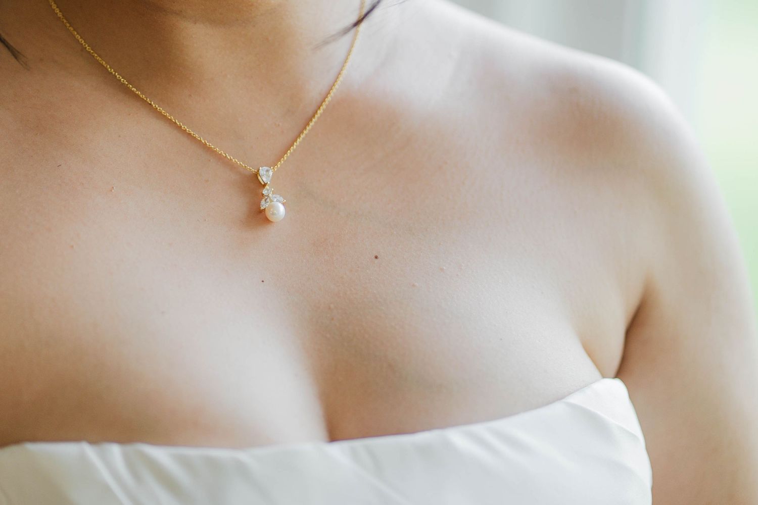 Close-up of a bride wearing a delicate gold necklace with pearl pendant over a white strapless wedding gown.