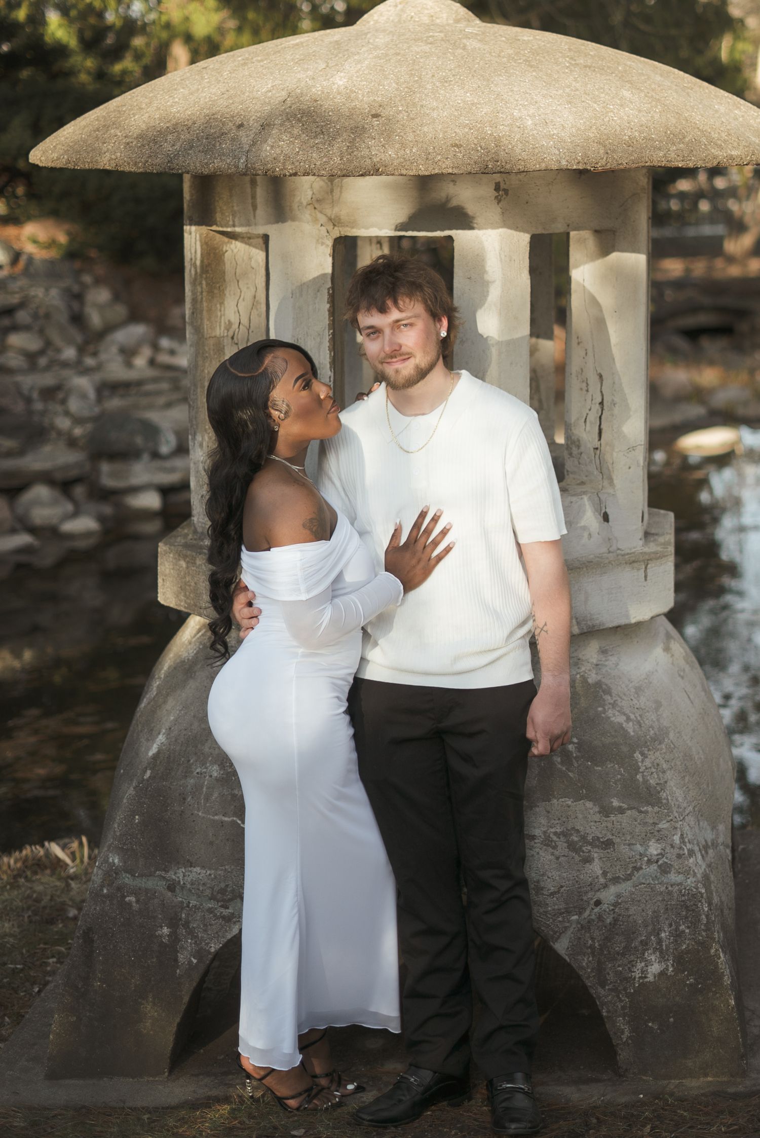 A couple poses romantically before a stone lantern, woman in white off-shoulder dress, man in white tee and black pants.