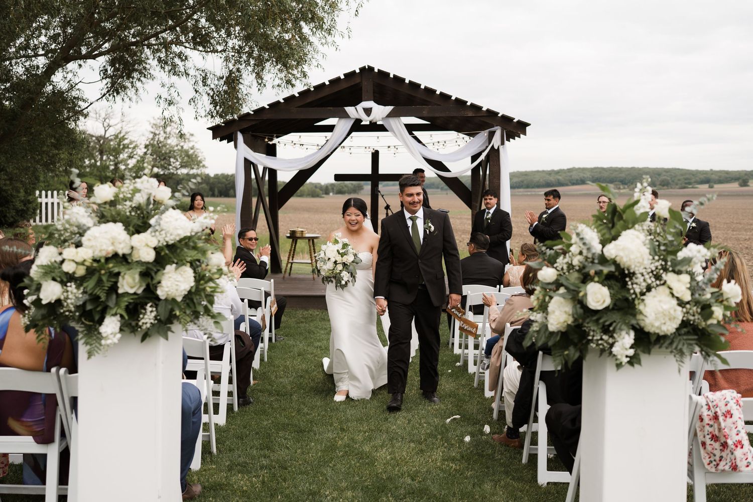 Newlyweds walk down aisle flanked by white floral arrangements at elegant outdoor wedding ceremony with rustic pergola backdrop.
