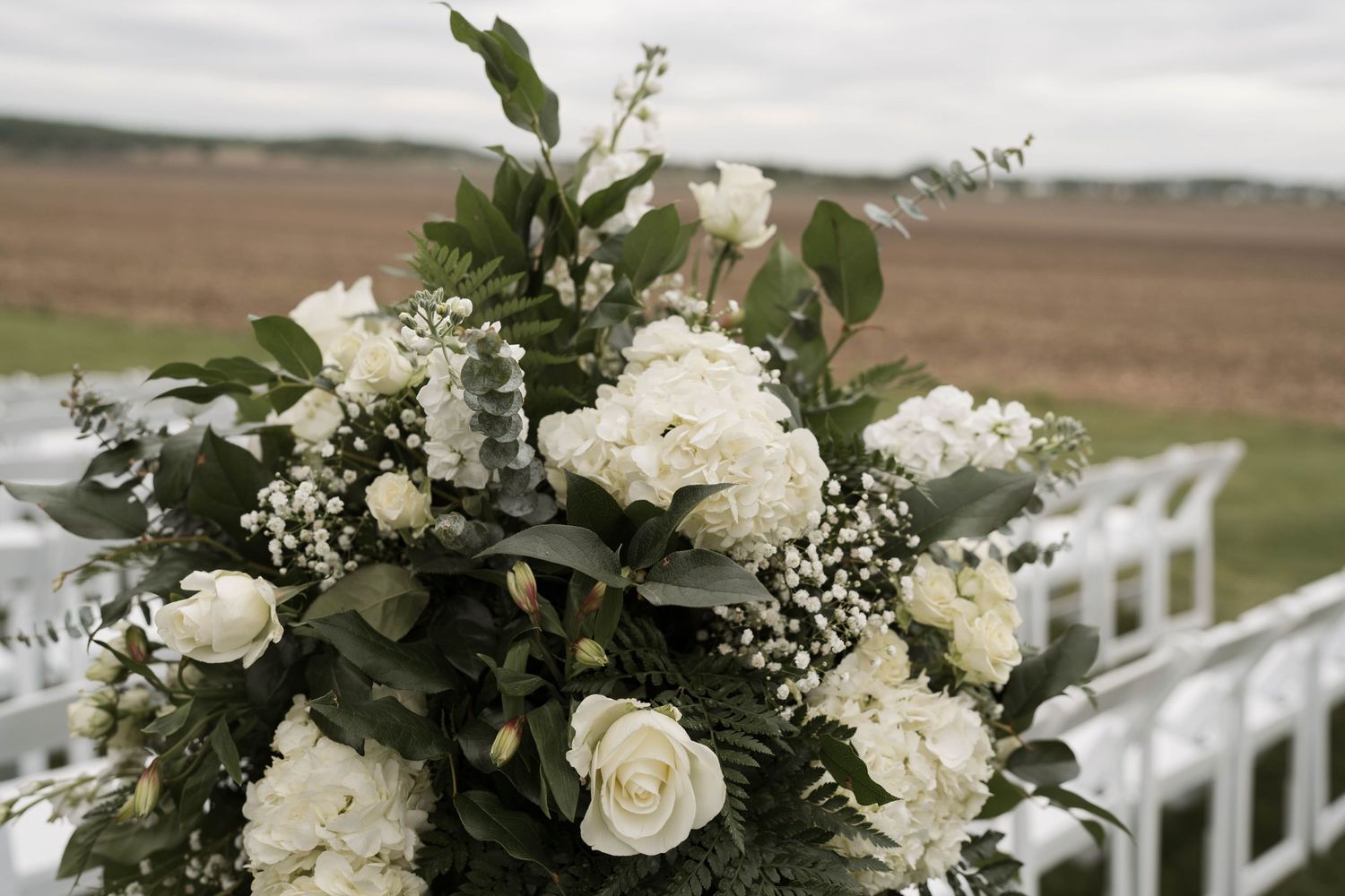 Elegant white wedding floral arrangement with hydrangeas, roses, and greenery at an outdoor ceremony.