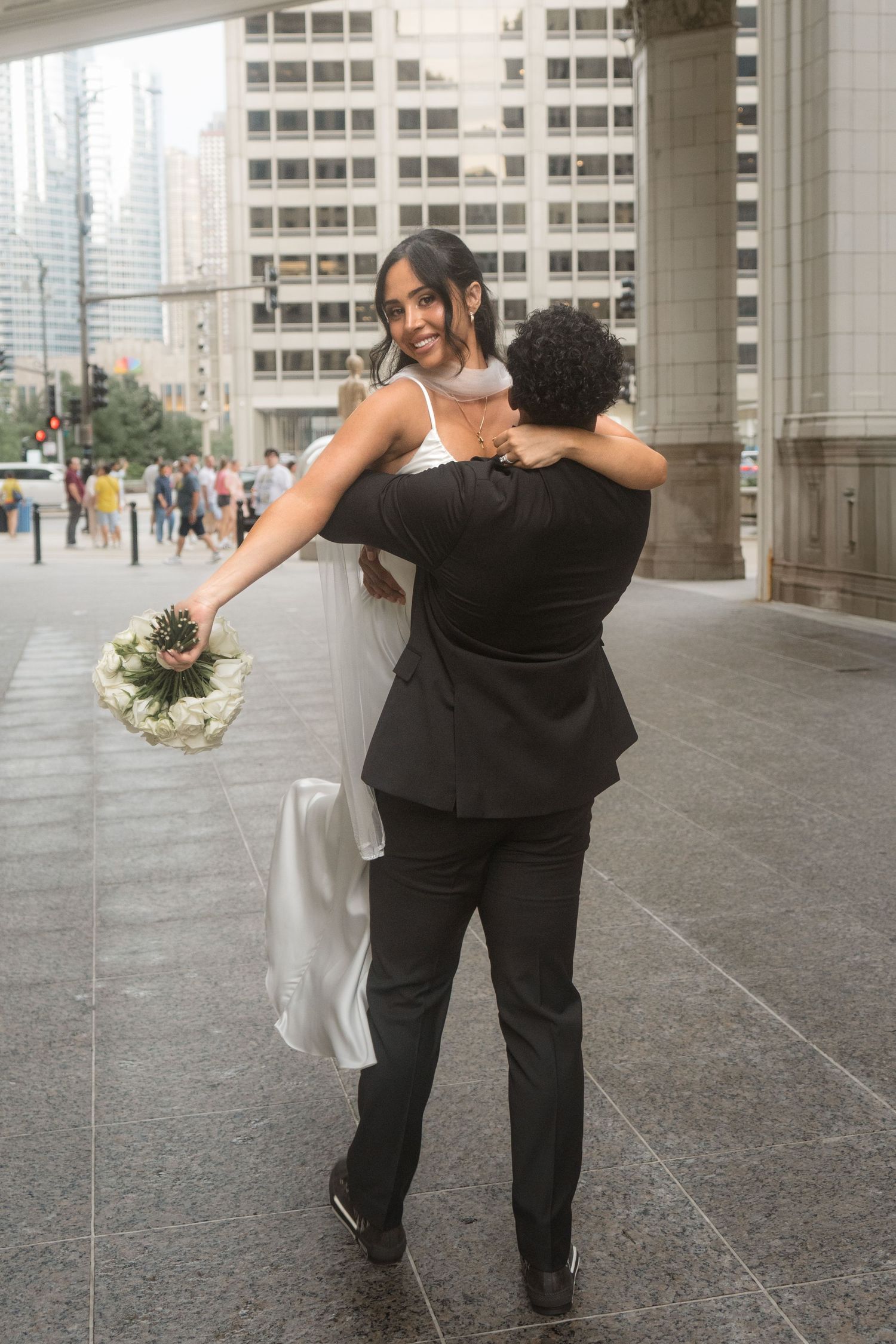 Groom lifting bride in white dress holding bouquet on urban city sidewalk during wedding celebration.