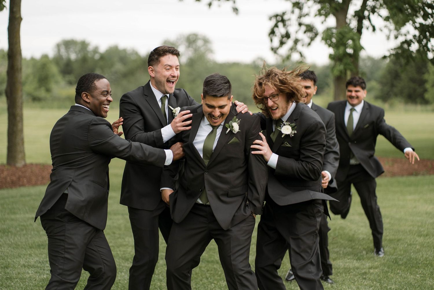 Groom surrounded by laughing groomsmen in dark suits with green ties celebrating outdoors at a wedding.