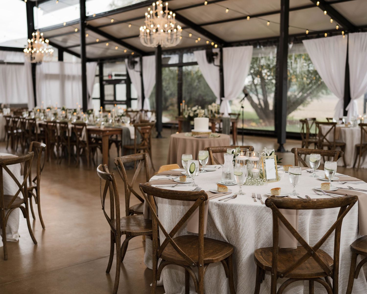 Elegant wedding reception venue with rustic wooden cross-back chairs, round tables, white linens, and chandelier lighting.
