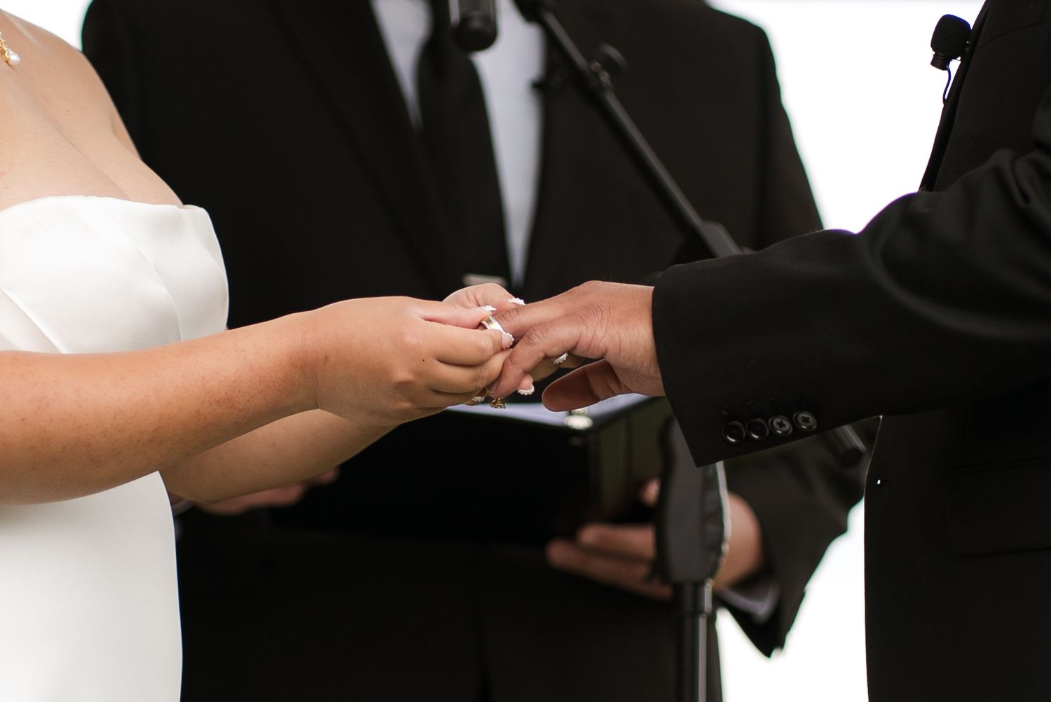 Bride placing wedding ring on groom's finger during ceremony with officiant holding book in background.