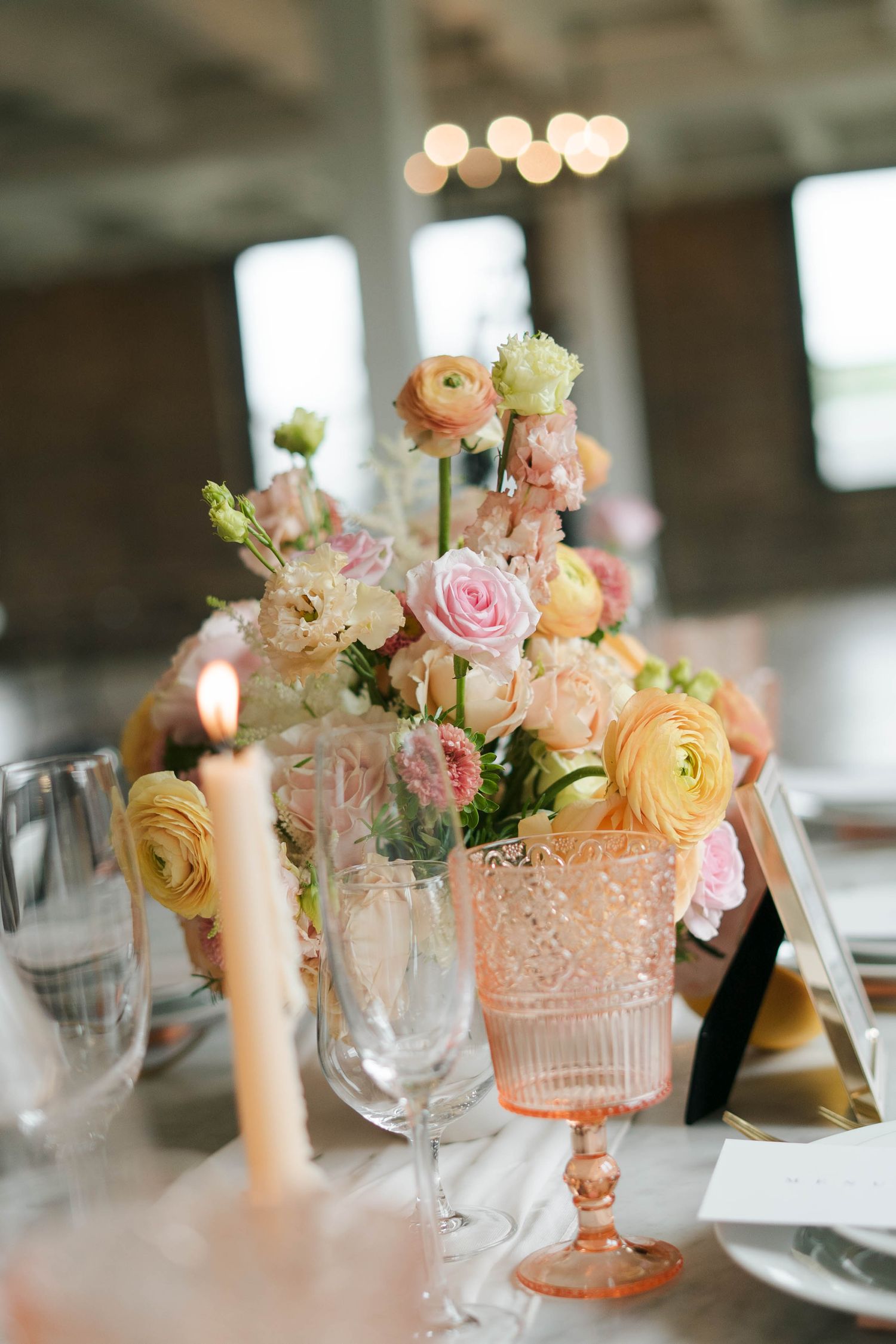 Elegant wedding table centerpiece with pink, peach, and yellow ranunculus roses in vintage glassware with lit taper candles.