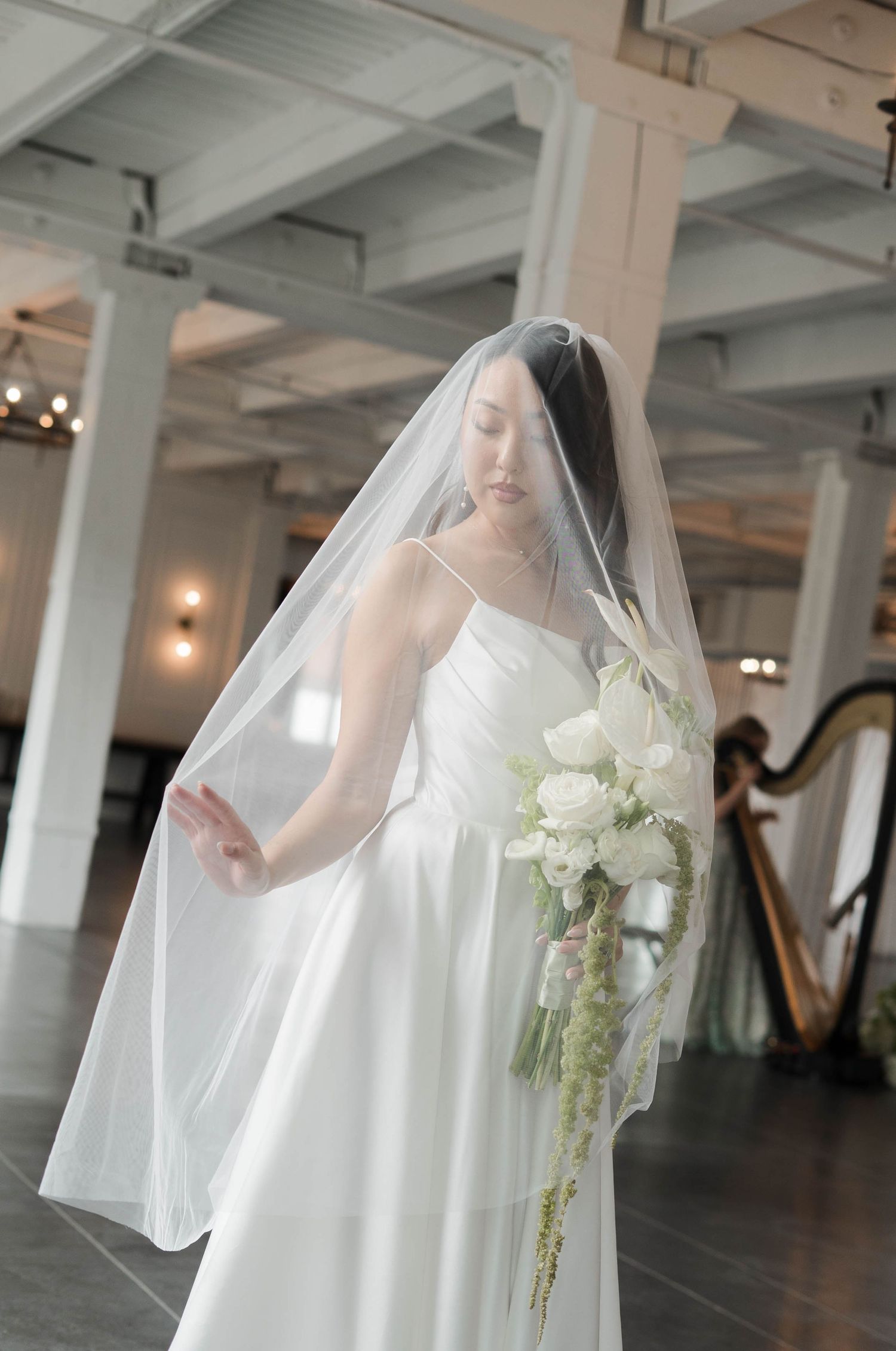 Bride in white gown and veil holds cascading white floral bouquet in elegant indoor venue with harp in background.