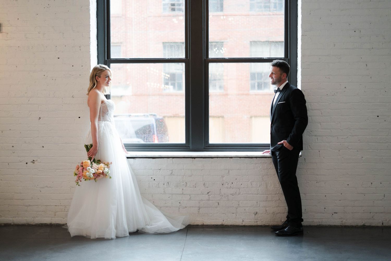 Bride in white gown holding colorful bouquet faces groom in black tuxedo near large windows in brick venue.