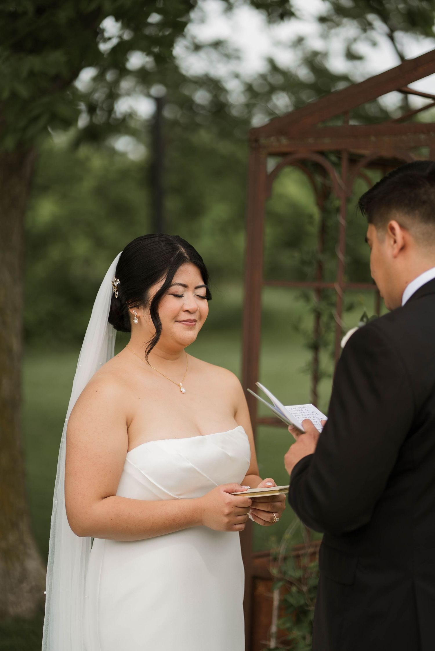 Bride in white strapless gown and veil reading wedding vows outdoors at a garden ceremony.