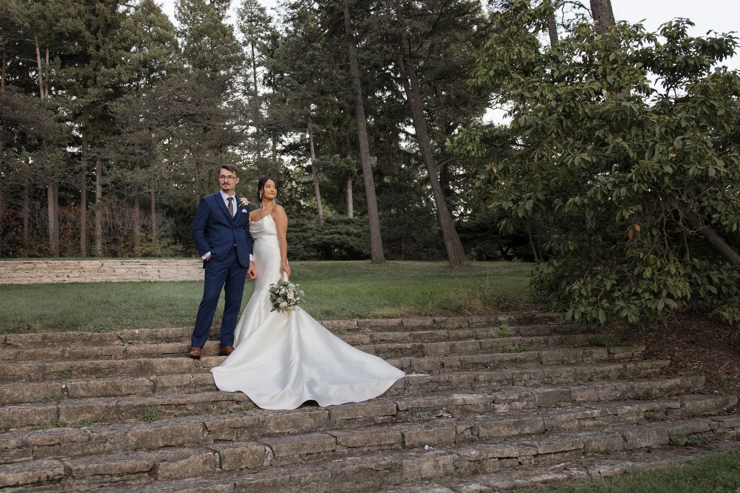 Bride and groom standing on stone steps surrounded by tall trees in an outdoor wedding venue.