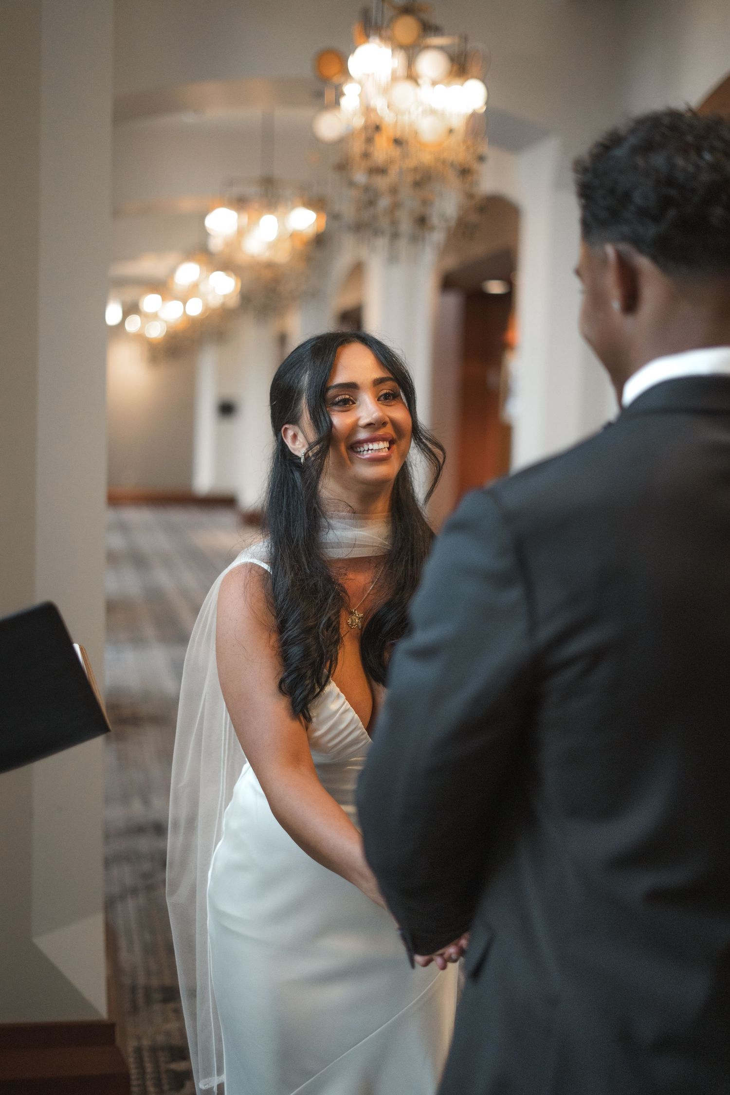 Smiling bride in white gown and veil holds hands with groom in black suit under elegant chandeliers.