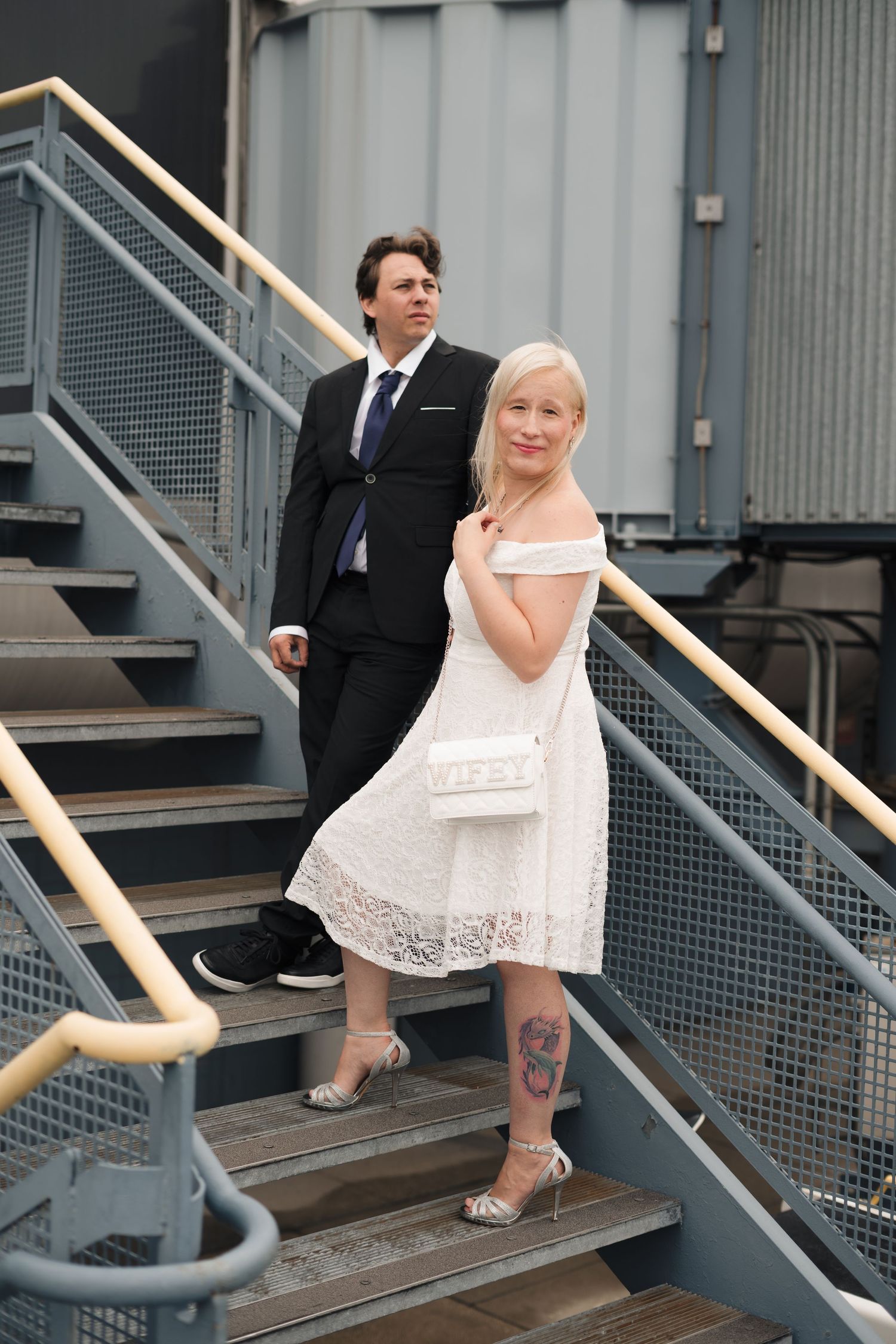 Couple in formal attire on industrial stairs, woman in white lace dress with tattoo, man in black suit.