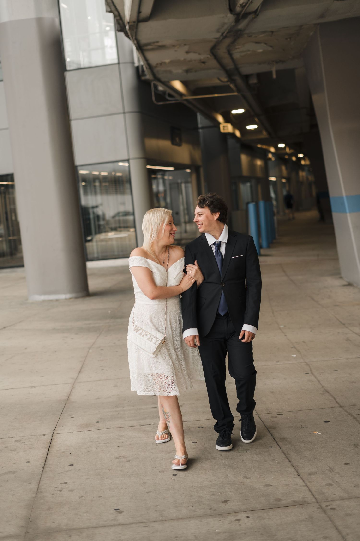 A couple in formal attire walks together outside a modern building, the woman in white dress, man in dark suit.