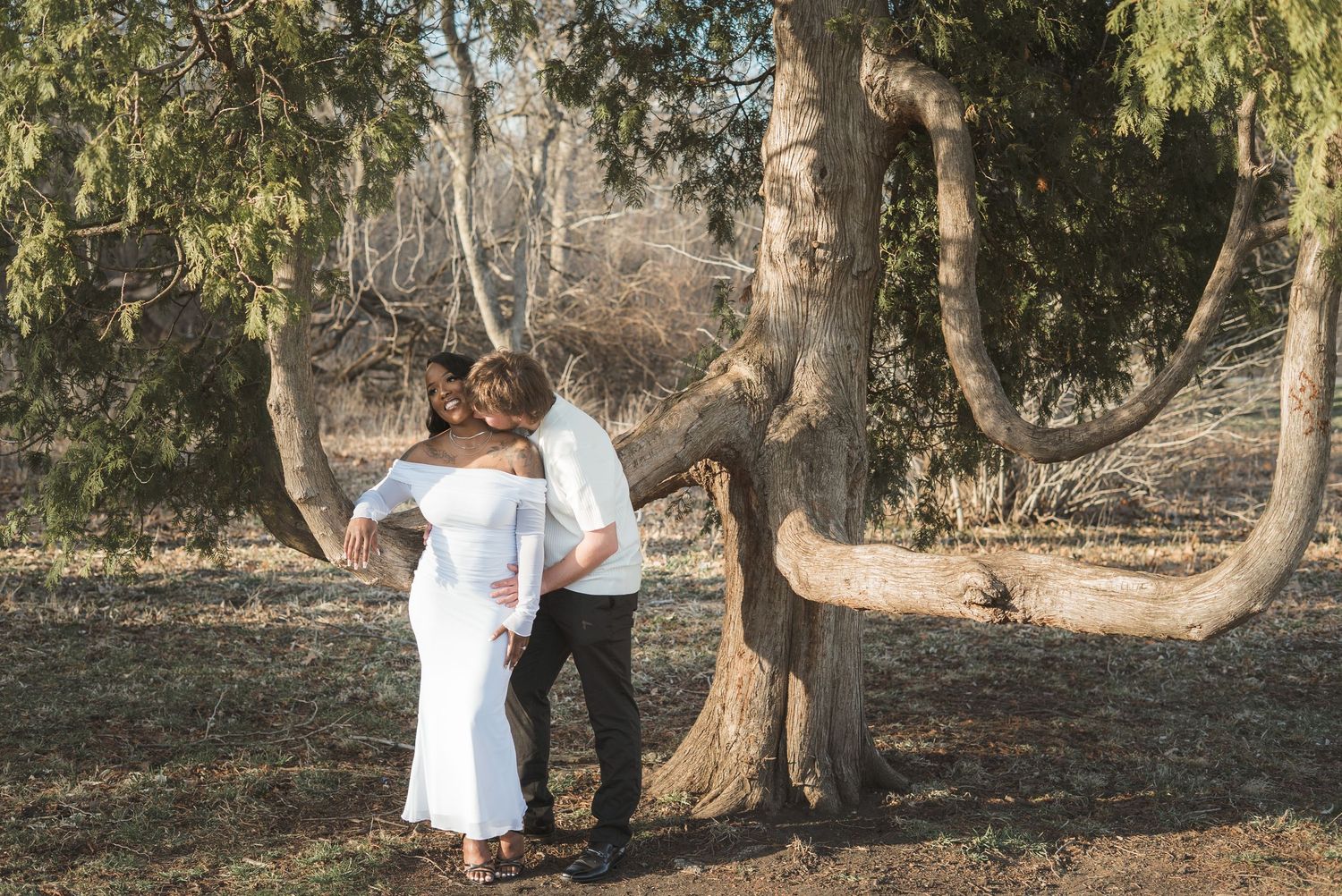 Couple embracing near a large tree in a park, woman in white off-shoulder dress, man in dark pants, outdoor maternity photo.
