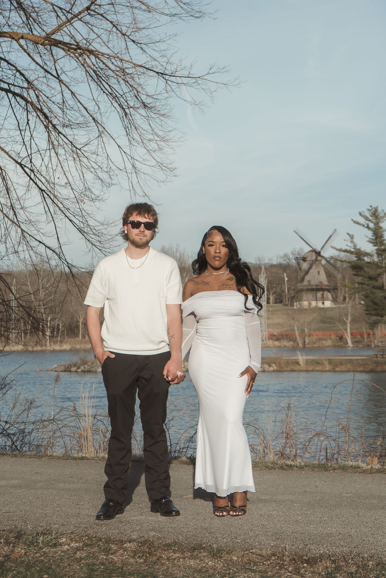 Couple holding hands in white dress and casual outfit by a lakeside with a windmill in the background.
