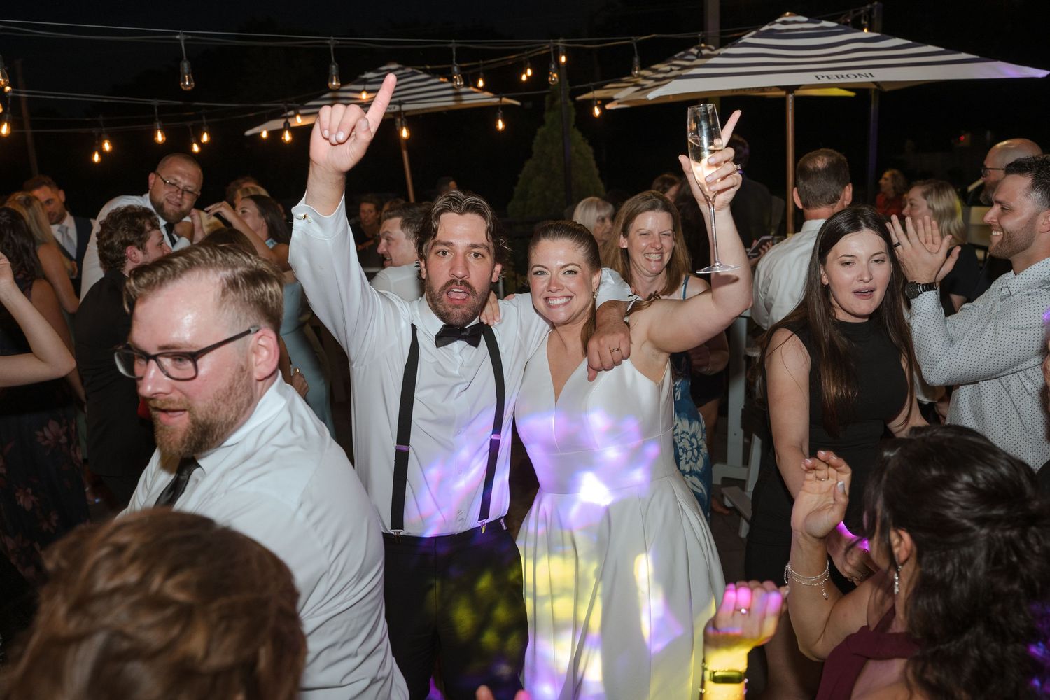 Bride and groom celebrate joyfully on the dance floor surrounded by wedding guests at an outdoor evening reception.