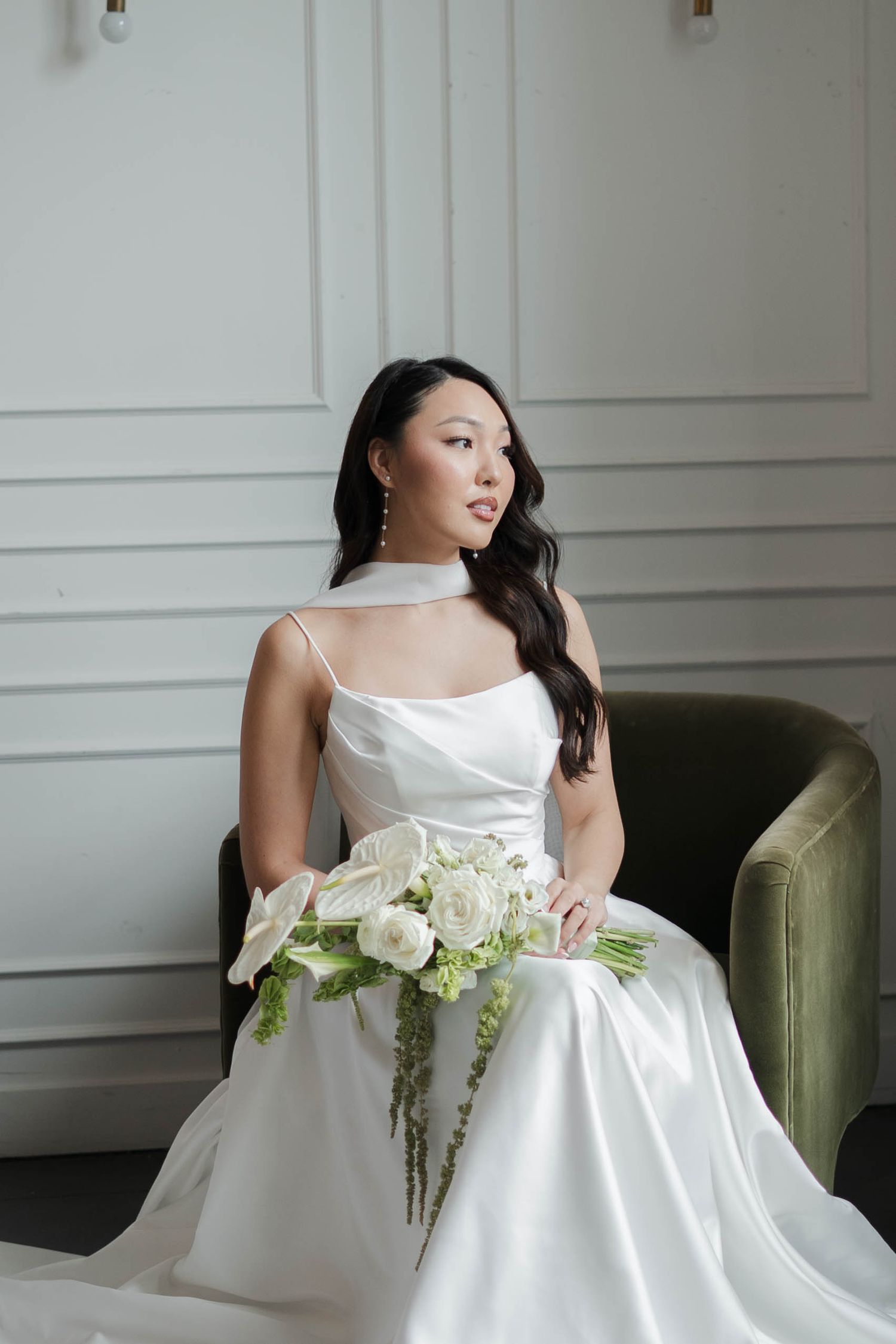 Bride in white satin gown holding white floral bouquet, seated in green velvet chair against elegant white paneled wall.