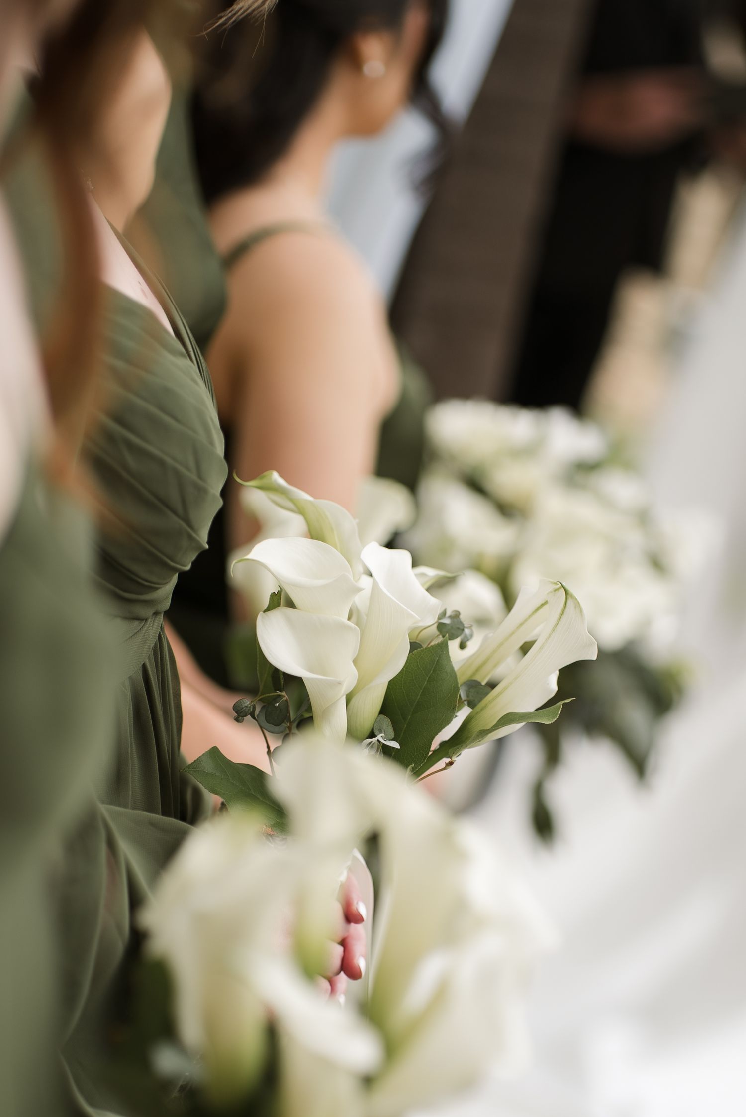 Bridesmaids in sage green dresses holding elegant white calla lily bouquets during a wedding ceremony.