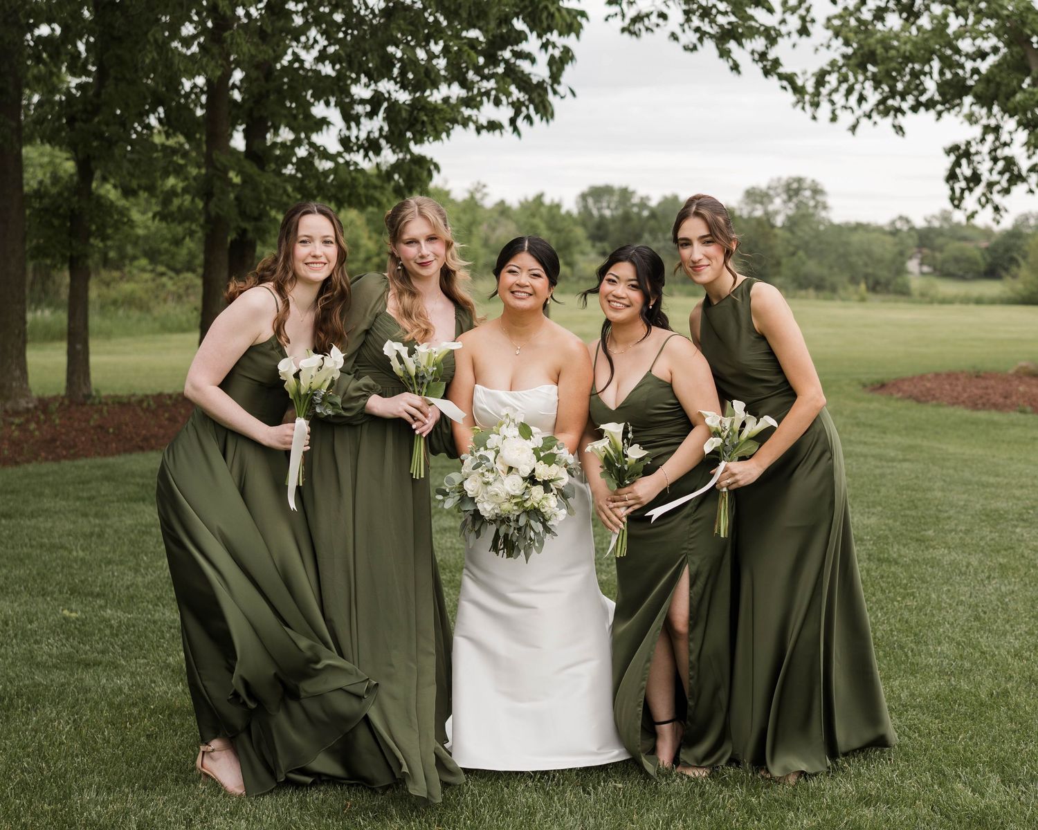 Bride in white gown surrounded by four bridesmaids in olive green dresses holding white bouquets outdoors.