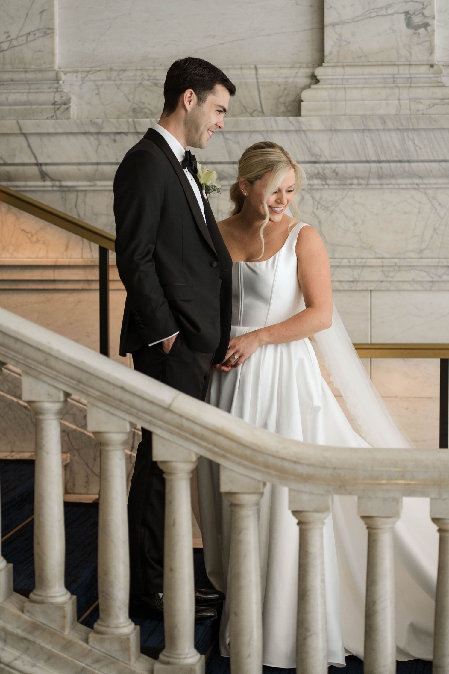 A bride and groom share a joyful moment on a grand marble staircase, she in a white gown, he in a black tuxedo.