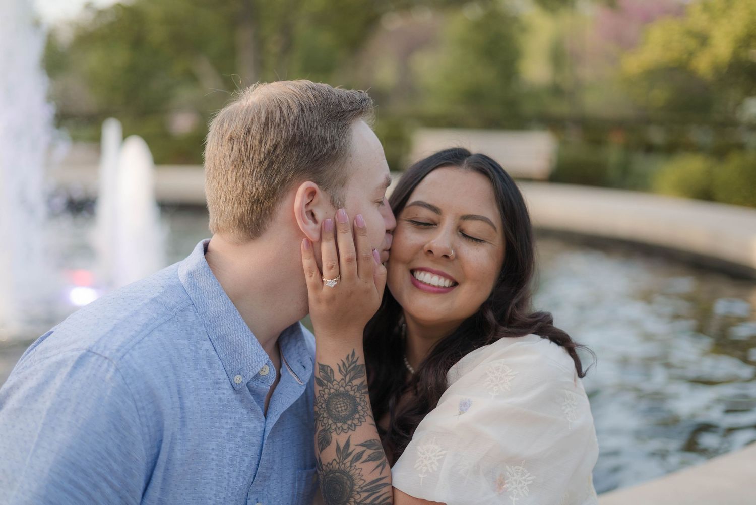 A man with a tattoo kisses a smiling woman on the cheek near a fountain outdoors.