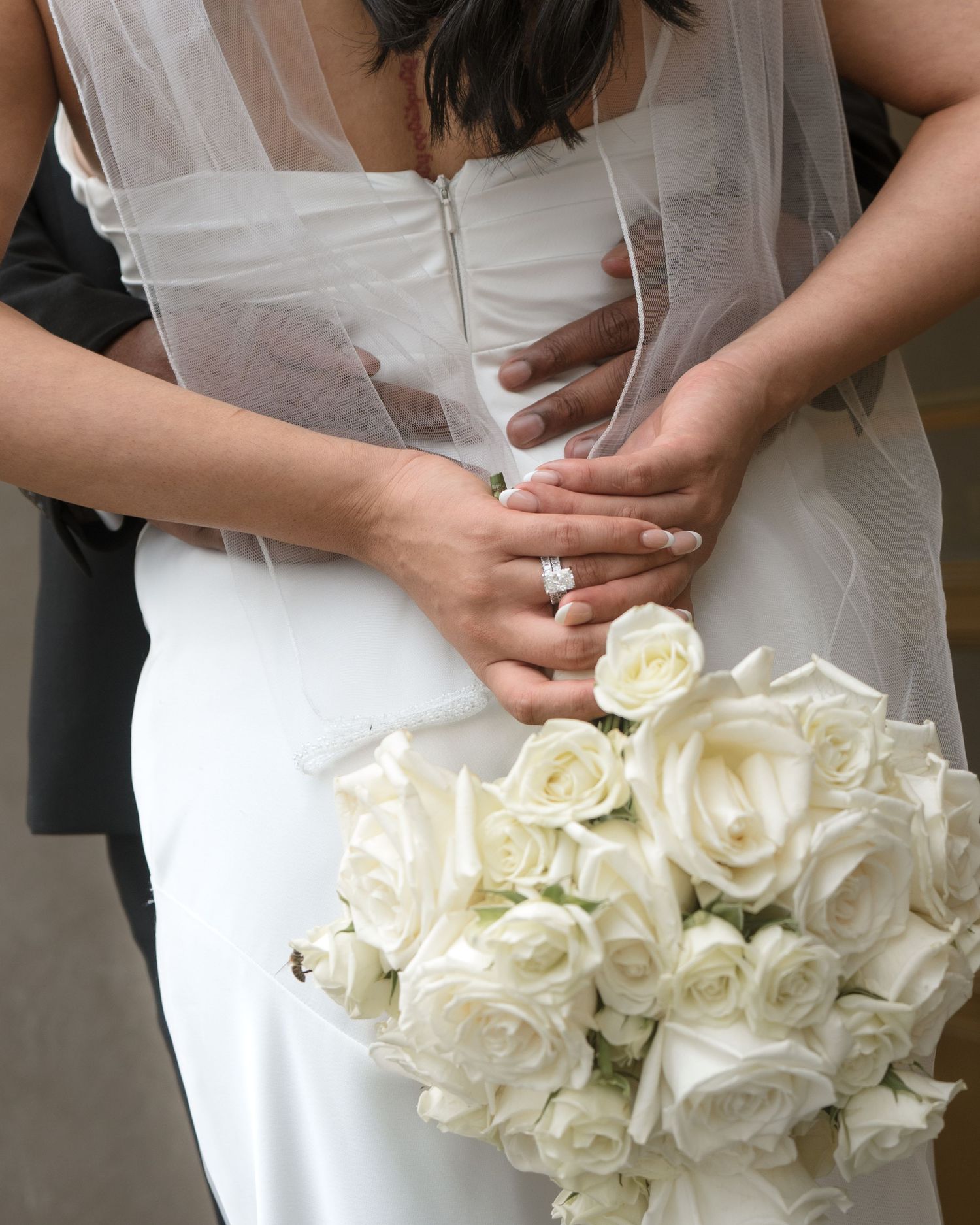 Bride holding white rose bouquet with diamond engagement ring visible, groom embracing her from behind.