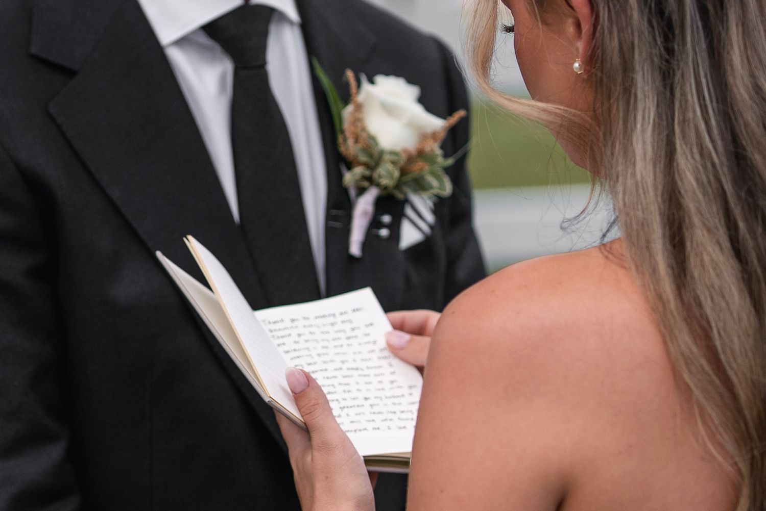 A couple reads handwritten vows during their wedding ceremony while standing close together outdoors.