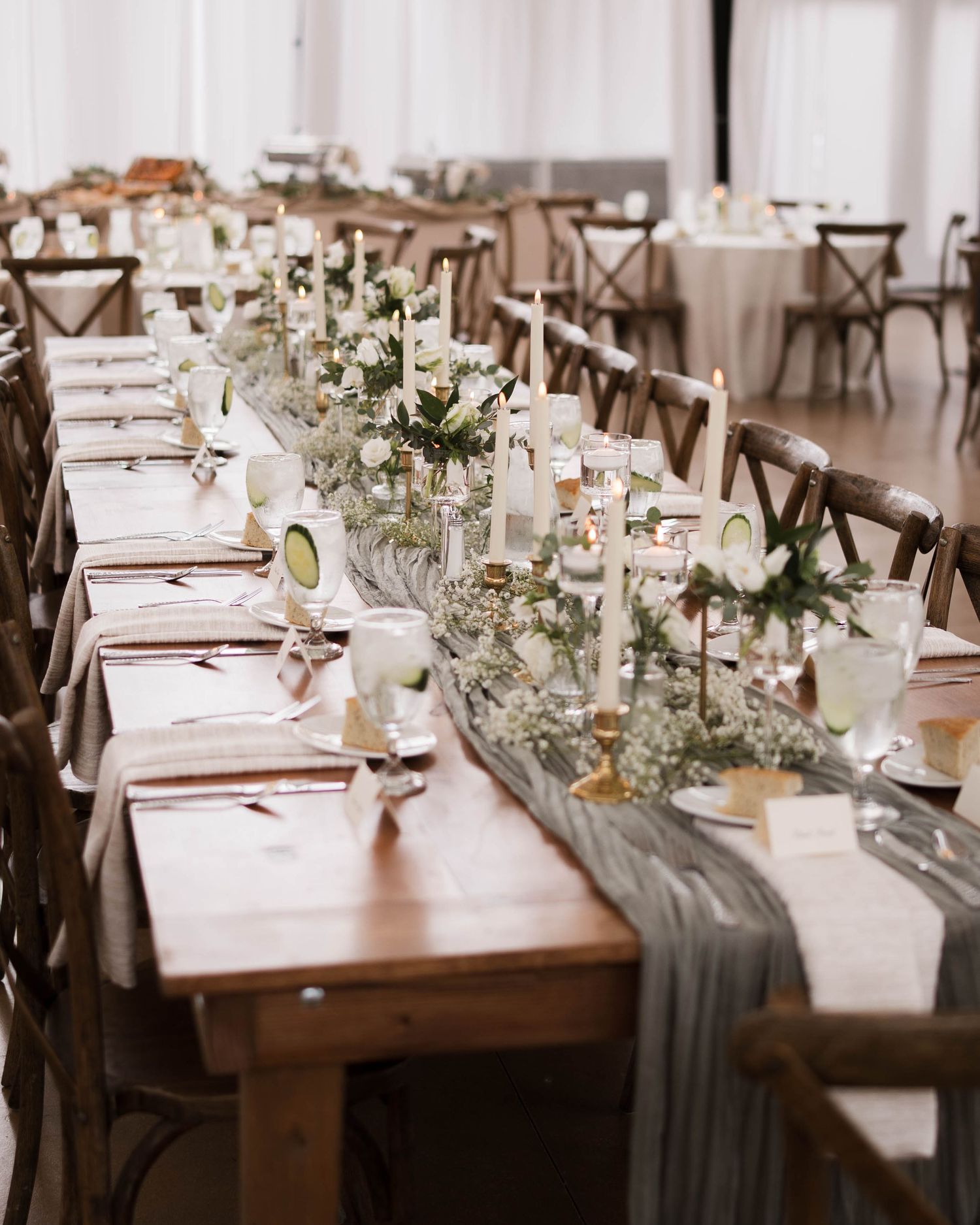 Rustic wedding reception table with wooden farm tables, sage green runner, white floral centerpieces, and candles in elegant setting.
